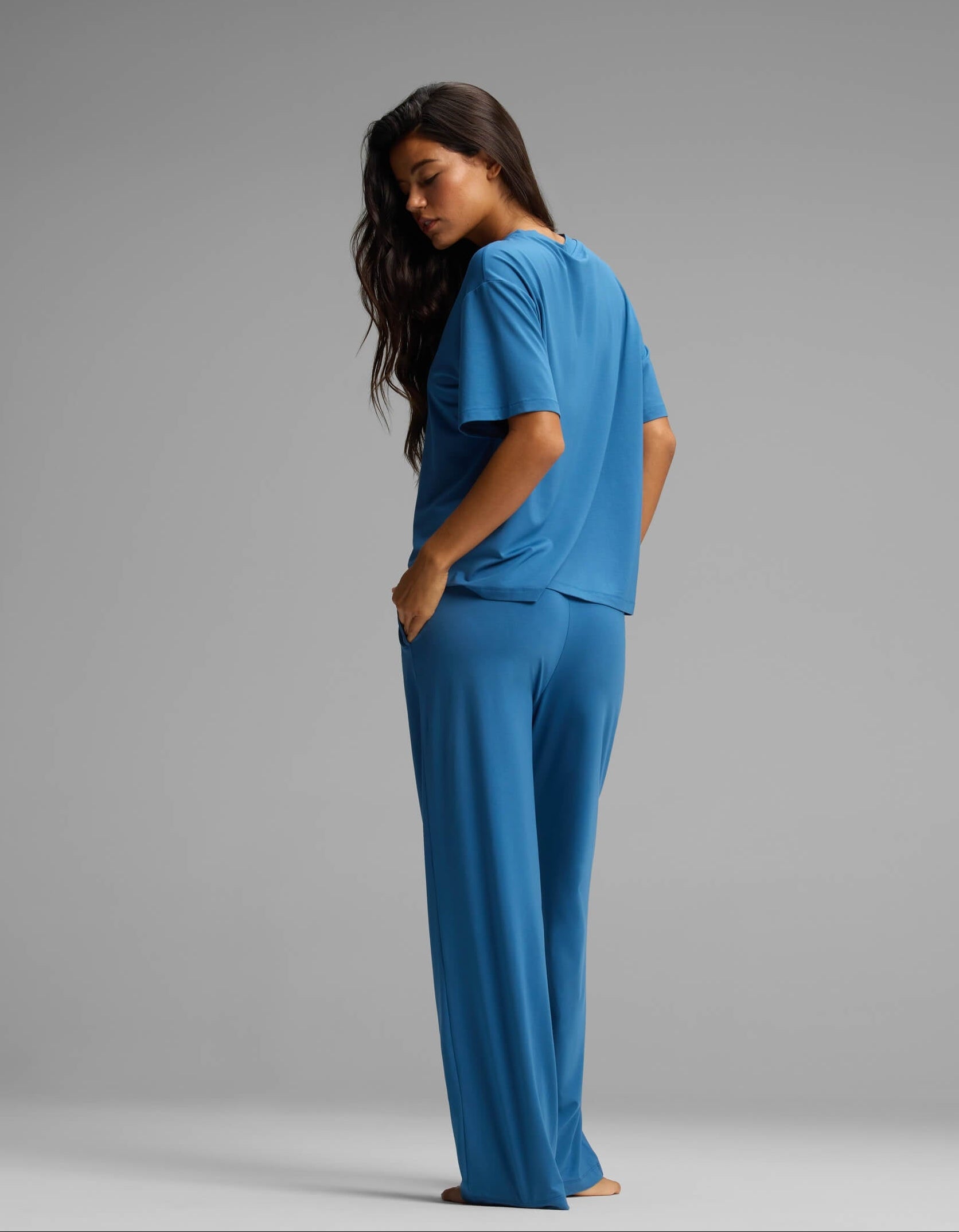 A woman stands barefoot, facing sideways and looking over her shoulder. She is wearing the Cozy Earth Women’s First Layer Pajama Short Sleeve Tee & Pant Set in blue against a plain grey background.