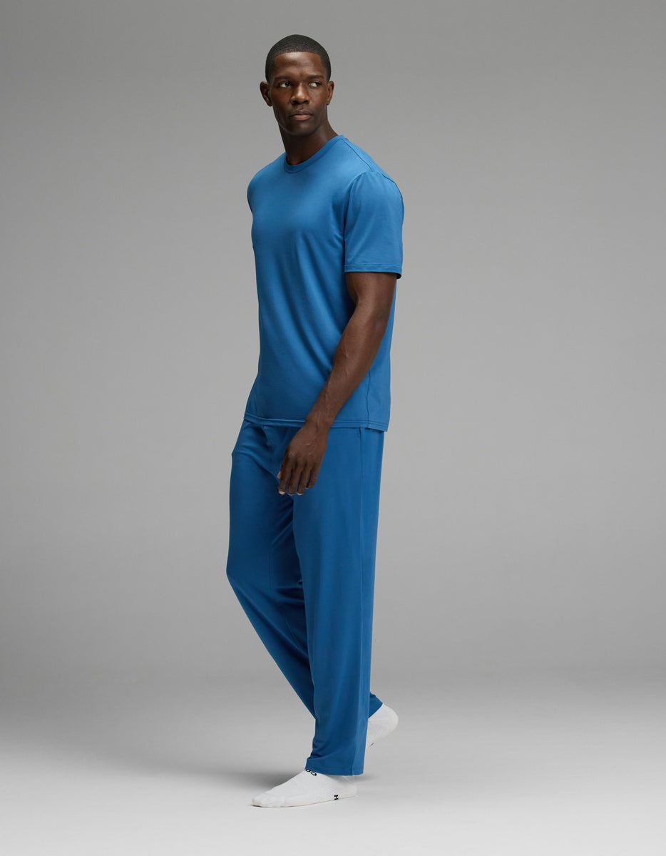 A man stands in a relaxed pose wearing the Cozy Earth Men’s First Layer Pajama Short Sleeve Tee & Pant Set in blue, paired with white shoes, against a plain gray background.