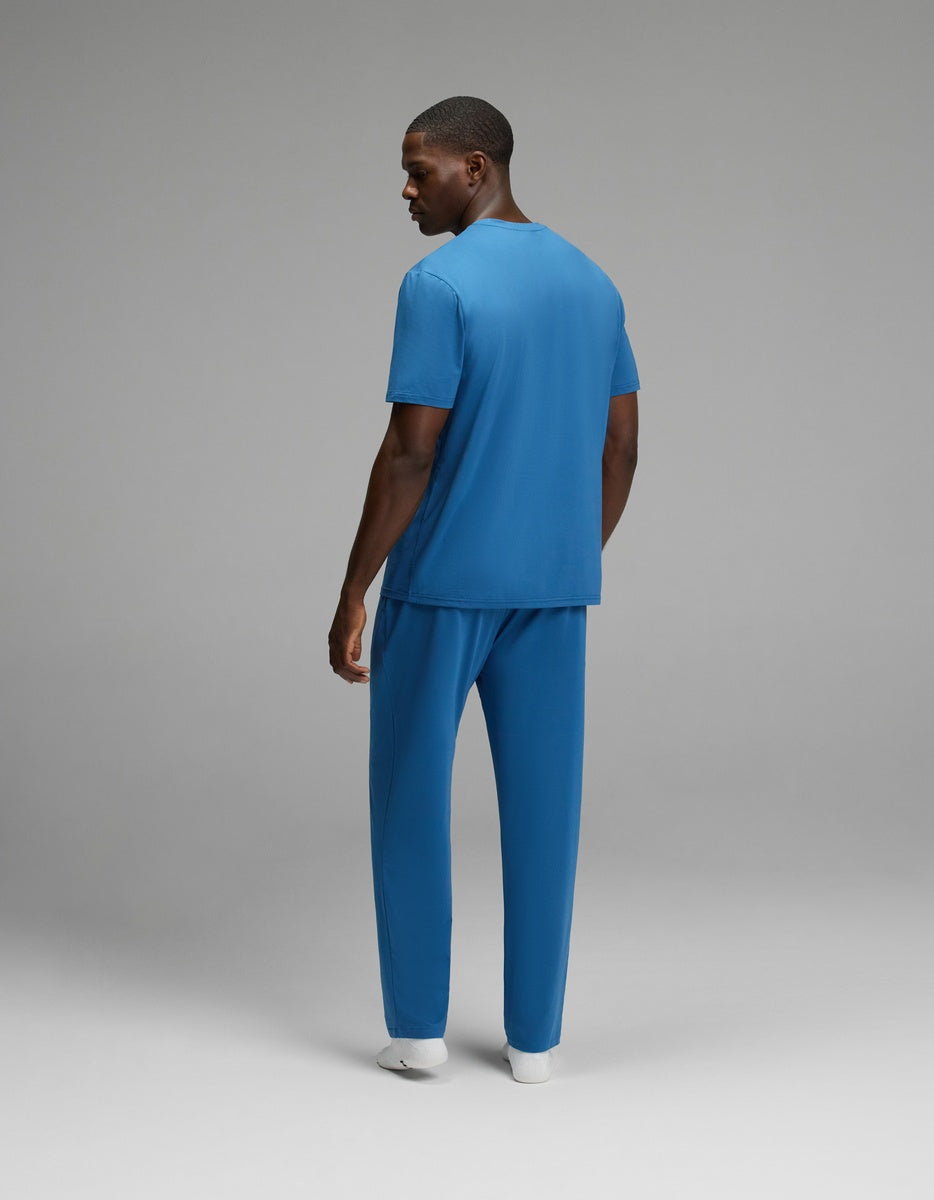 A man models the Cozy Earth Men’s First Layer Pajama Short Sleeve Tee & Pant Set in blue, standing slightly turned with a glance over his shoulder against a plain gray background.