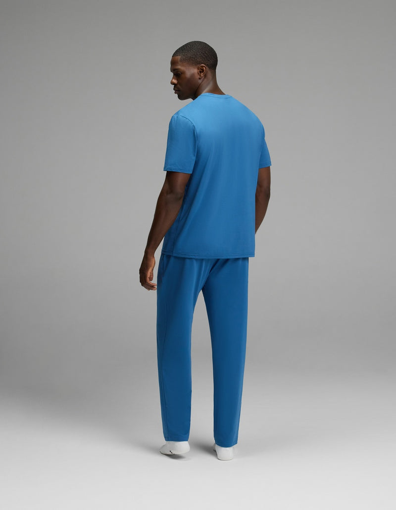 A man models the Cozy Earth Men’s First Layer Pajama Short Sleeve Tee & Pant Set in blue, standing slightly turned with a glance over his shoulder against a plain gray background.