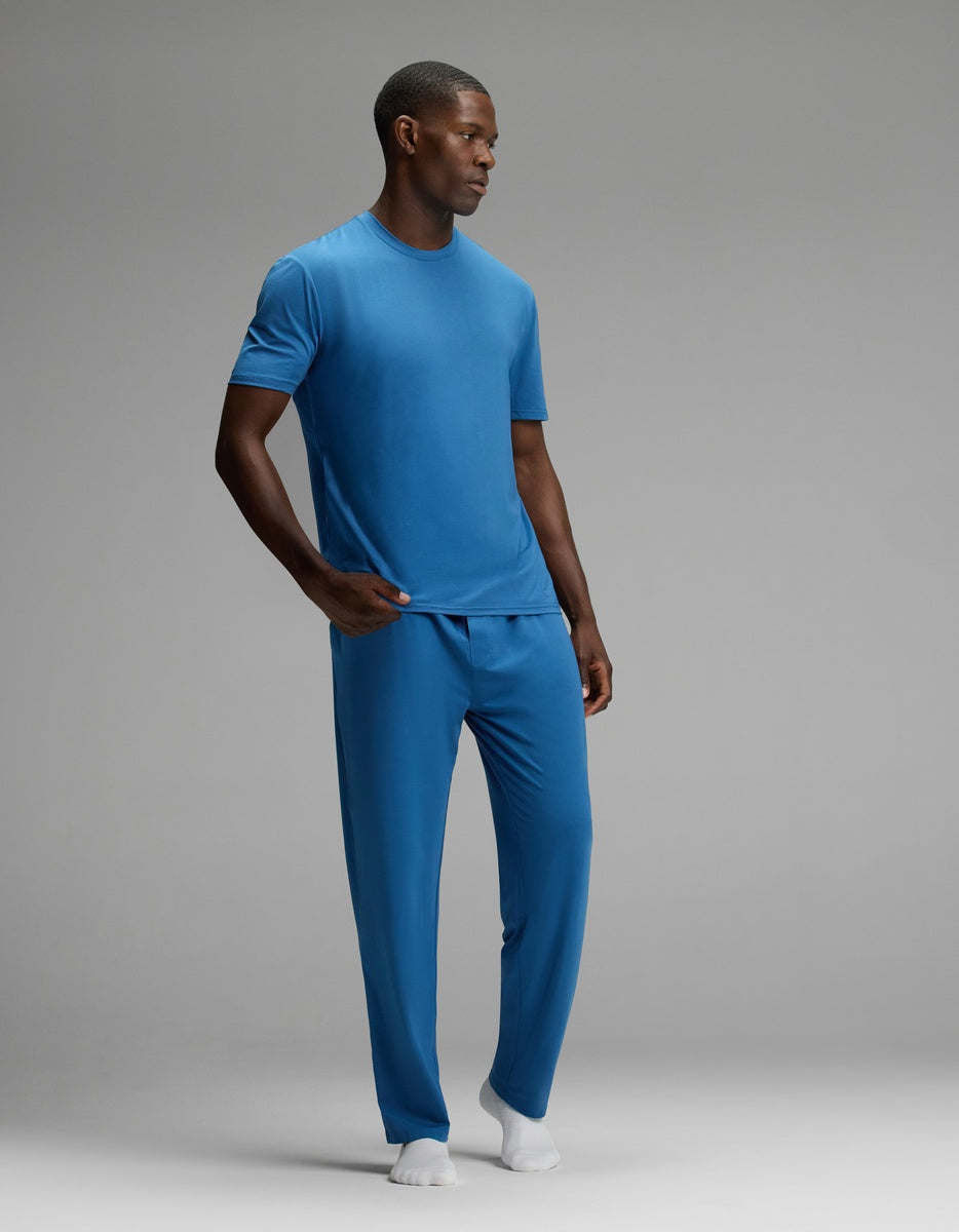 A man stands in a studio wearing the Cozy Earth Men’s First Layer Pajama Short Sleeve Tee & Pant Set in light blue with white socks. The background is plain gray, and he is looking to the side with one hand in his pocket.