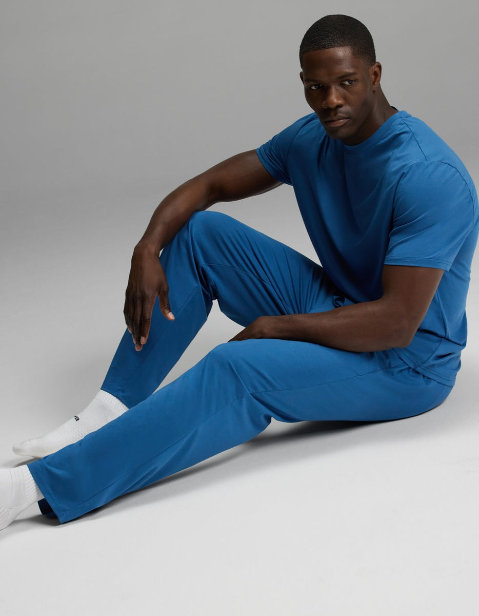 A man wearing the Cozy Earth Men’s First Layer Pajama Short Sleeve Tee & Pant Set in blue sits on the floor with one knee up and the other leg extended, looking toward the camera against a plain, neutral background.
