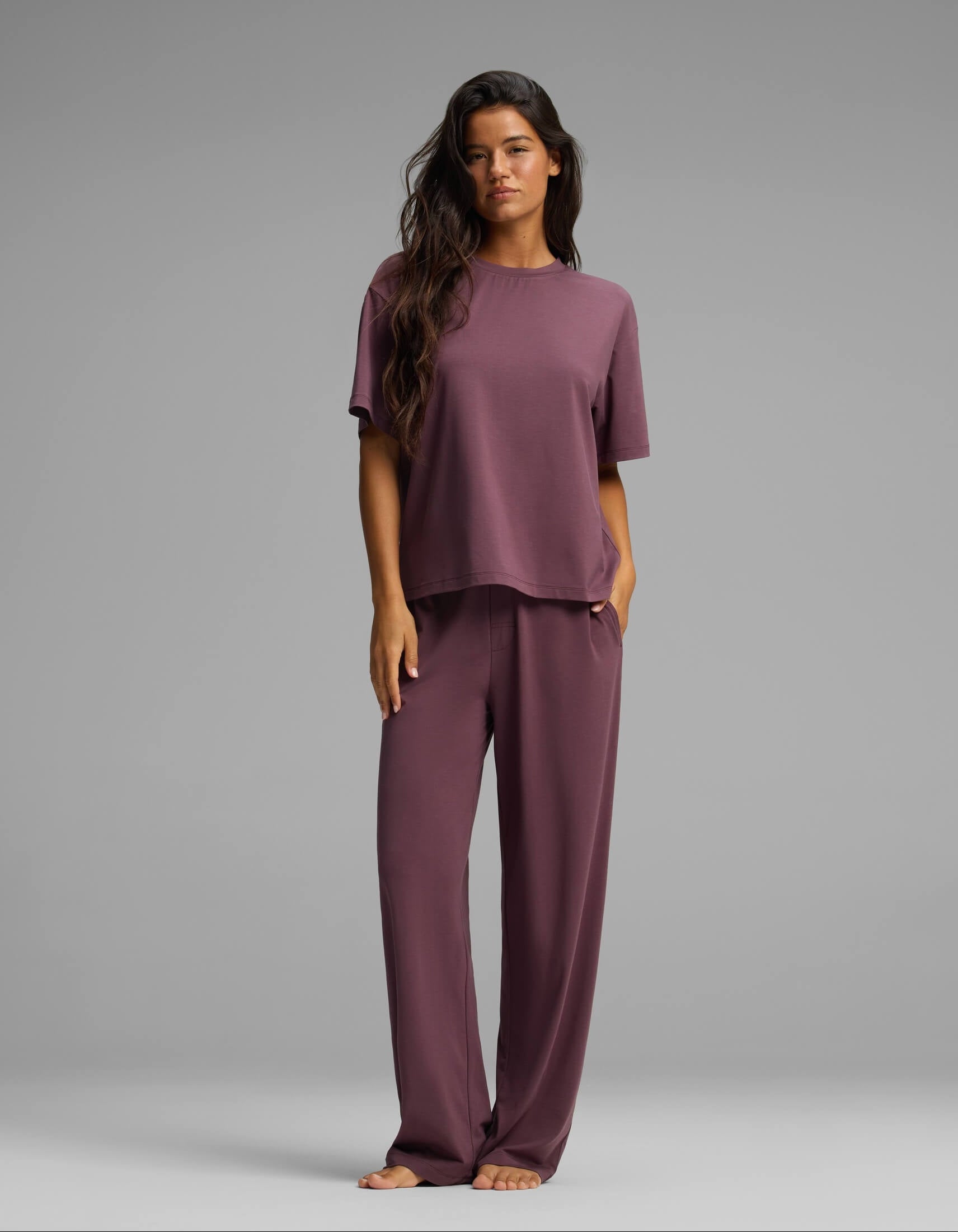 A woman with long dark hair is seen standing barefoot against a plain gray background, wearing the Cozy Earth Women’s First Layer Pajama Short Sleeve Tee & Pant Set in a loose-fitting purple style with matching wide-leg pants.