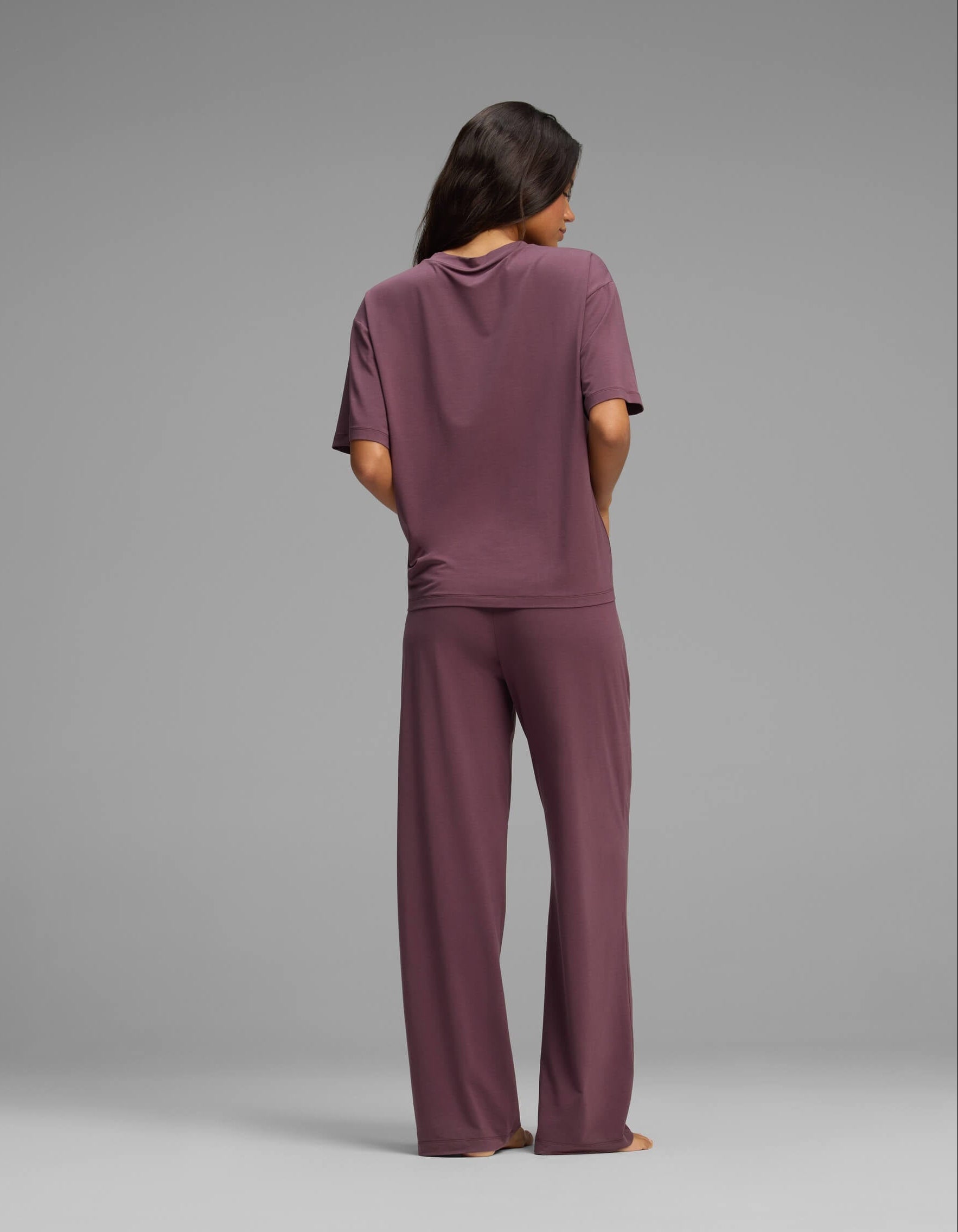 A person with long hair stands barefoot facing away, wearing the Cozy Earth Women’s First Layer Pajama Short Sleeve Tee & Pant Set in mauve against a plain gray background.