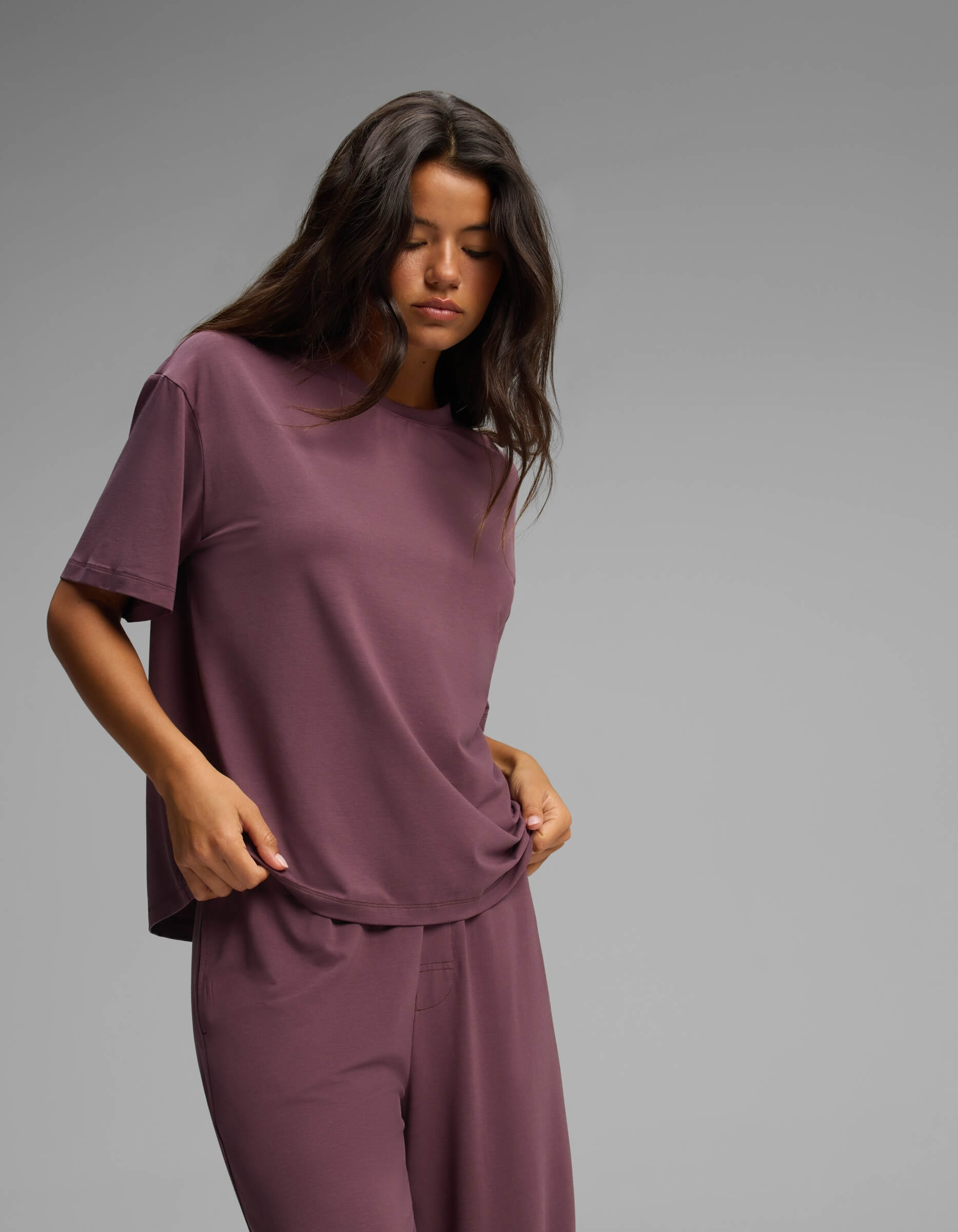A woman with long dark hair wears the Cozy Earth Women’s First Layer Pajama Short Sleeve Tee & Pant Set in purple. She stands against a plain gray background, looking down with a relaxed expression as she lightly holds the hem of her shirt.