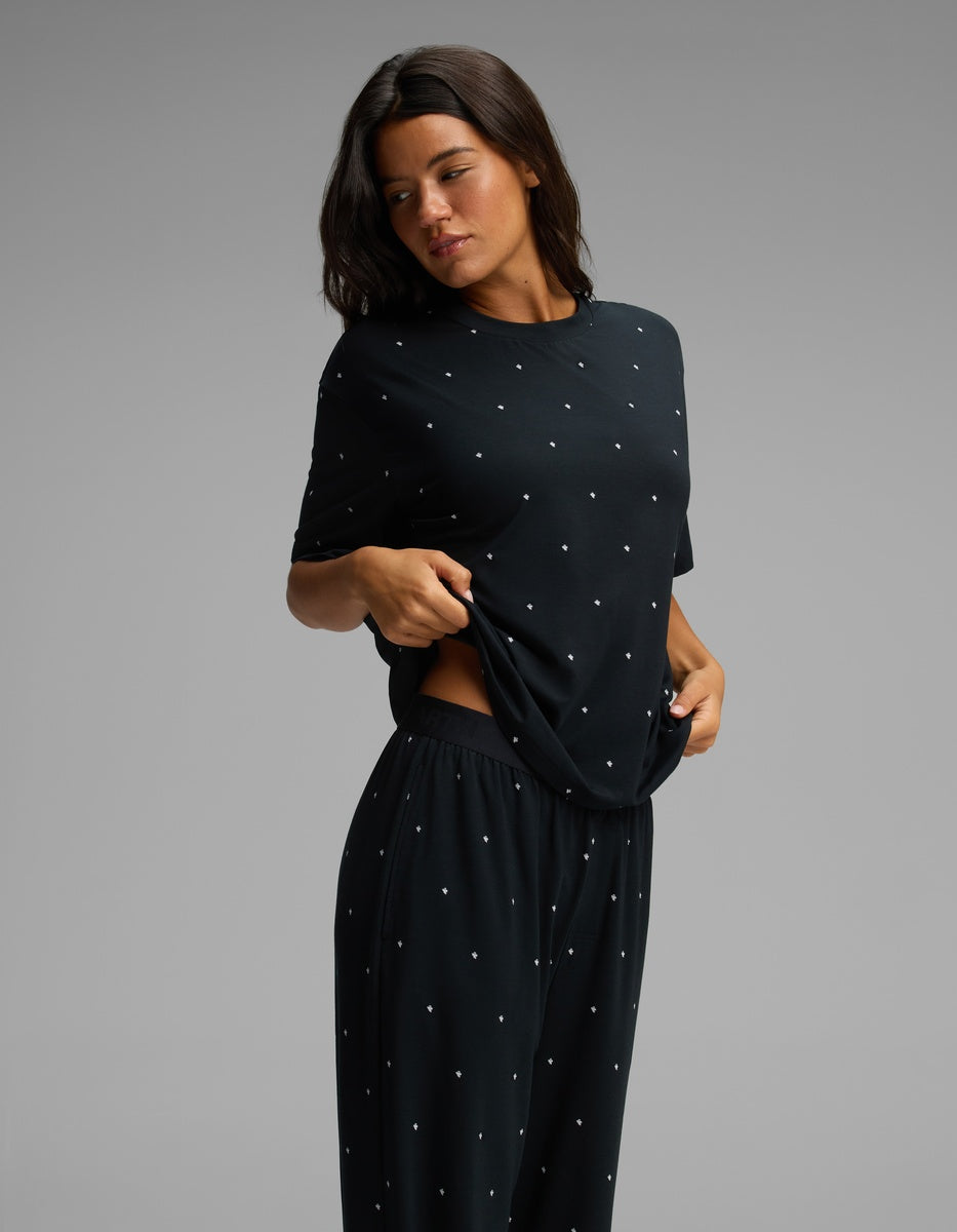 A woman wearing the Cozy Earth Women’s First Layer Pajama Short Sleeve Tee & Pajama Pant Set in black with small white dots lifts her shirt slightly and looks down, standing against a plain gray background.