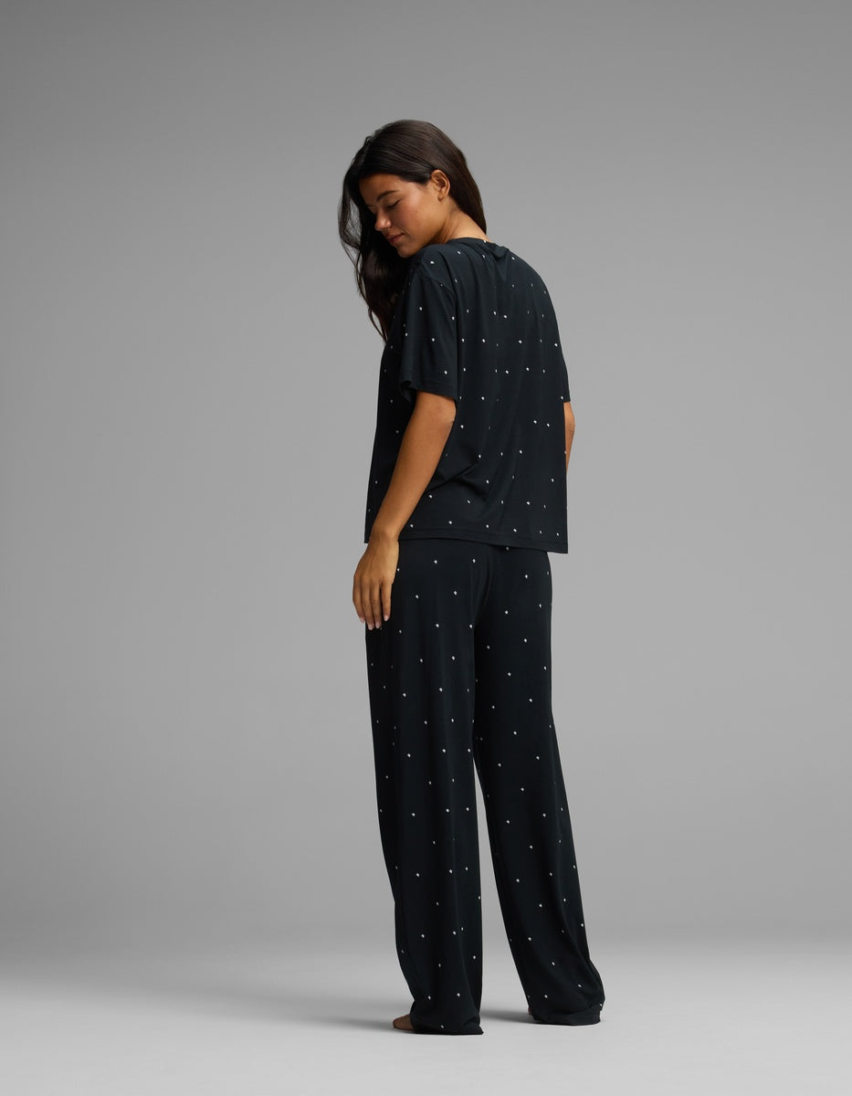 A woman with long dark hair stands barefoot, wearing the Cozy Earth Women’s First Layer Pajama Short Sleeve Tee & Pajama Pant Set in a loose fit with small white dots, looking over her shoulder against a plain gray background.