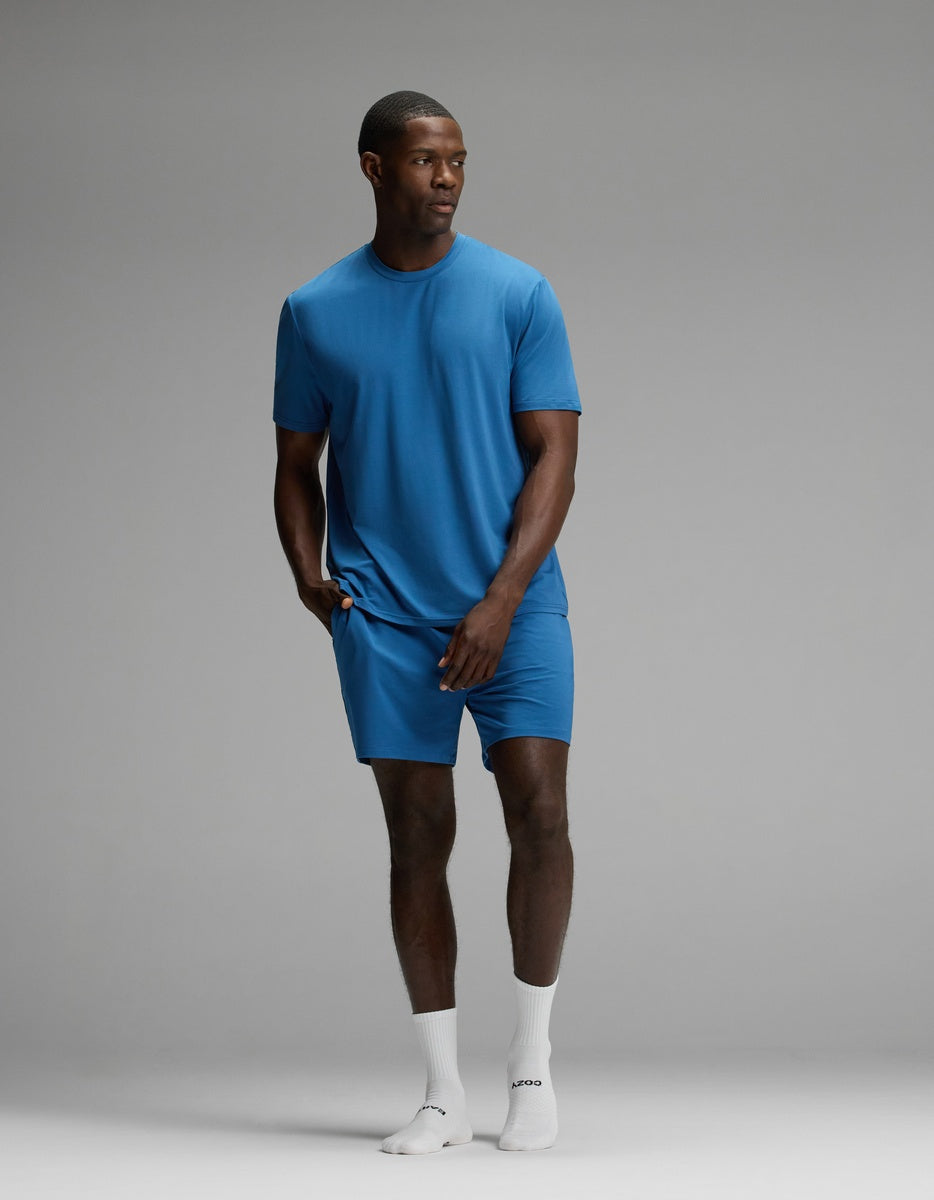 A man stands in a studio wearing the Cozy Earth Men’s First Layer Pajama Short Sleeve Tee & Short Set in blue, paired with white socks. The background is plain and gray, and he gazes off to the side with one hand casually resting in his shorts pocket.|Color:Anchor