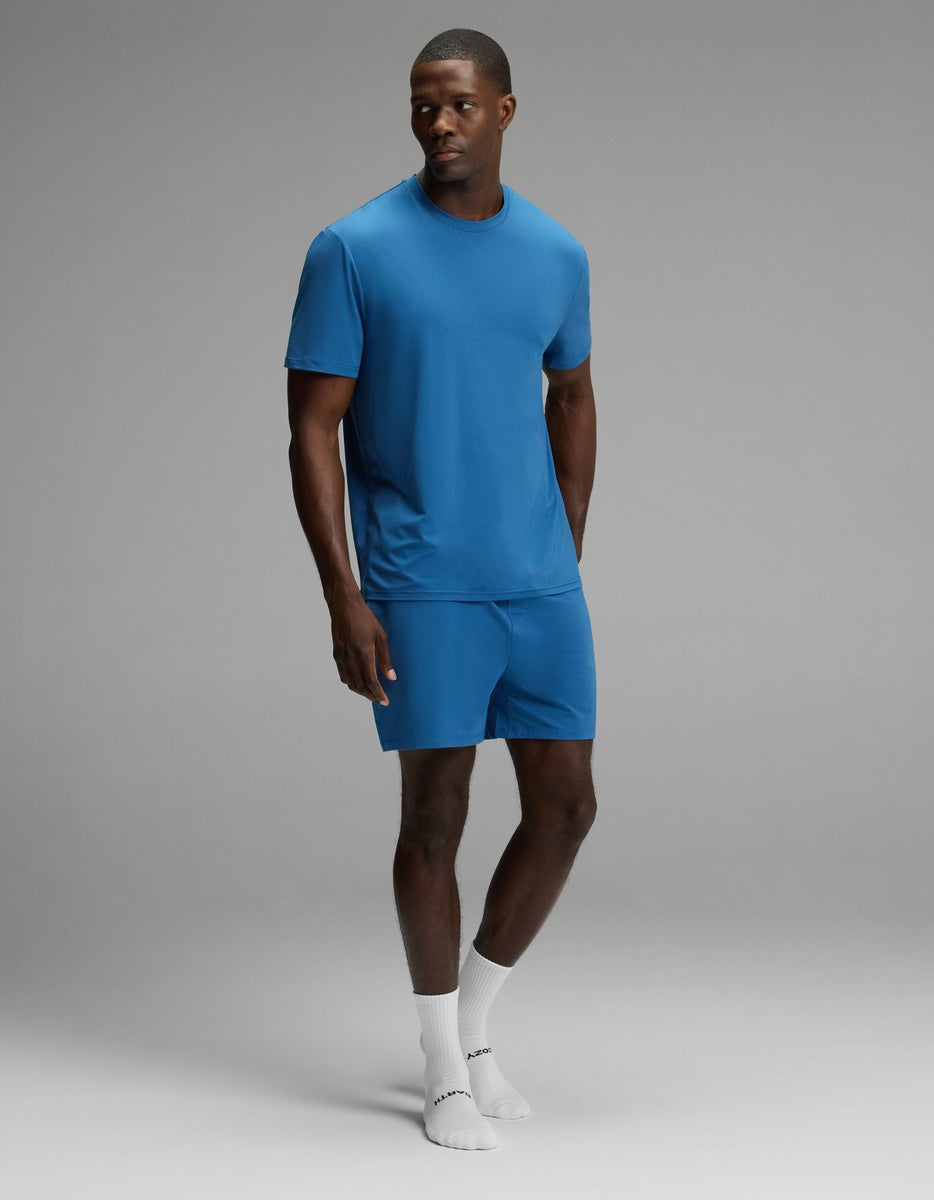 A man stands against a plain gray background wearing the Cozy Earth Men’s First Layer Pajama Short Sleeve Tee & Short Set in blue, paired with white socks. He looks slightly to the side with one hand in his pocket.