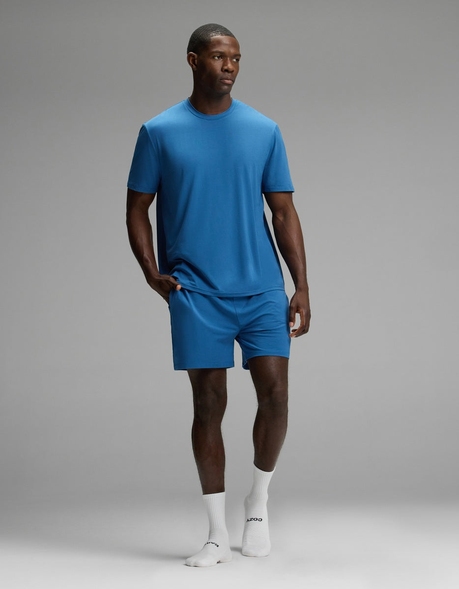 A man stands against a grey background, wearing the Cozy Earth Men’s First Layer Pajama Short Sleeve Tee & Short Set in blue with white crew socks. He has one hand in his pocket and looks slightly to the side.