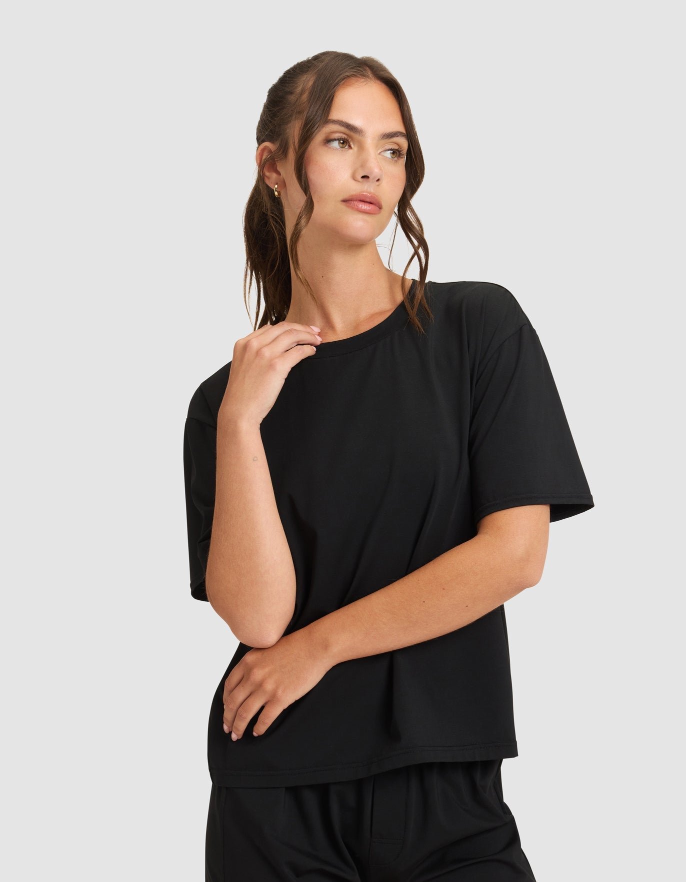 A woman wears the Cozy Earth Women's First Layer Pajama Short Sleeve Tee and matching shorts in black, standing against a light gray background, gazing to the side with one hand near her face and the other on her arm.|Color:Jet Black