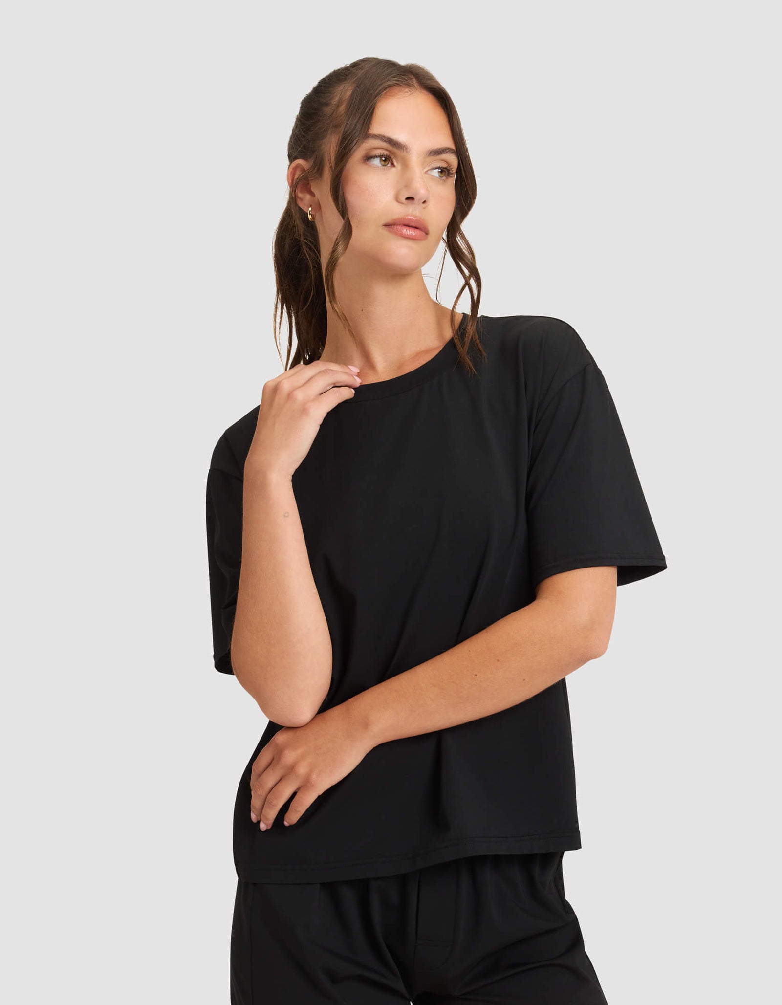 A woman wears the Cozy Earth Women's First Layer Pajama Short Sleeve Tee and matching shorts in black, standing against a light gray background, gazing to the side with one hand near her face and the other on her arm.
