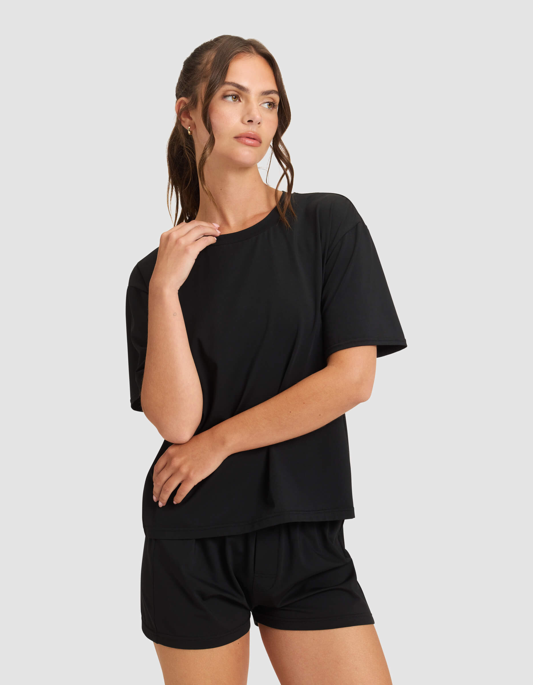 A woman wears the Cozy Earth Women's First Layer Pajama Short Sleeve Tee and matching shorts in black, standing against a light gray background, gazing to the side with one hand near her face and the other on her arm.|Color:Jet Black