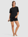 A woman stands barefoot against a plain background, wearing the Cozy Earth Women's First Layer Pajama Short Sleeve Tee and black shorts. She holds a reusable cup in her right hand, looking relaxed and smiling gently.|Color:Jet Black