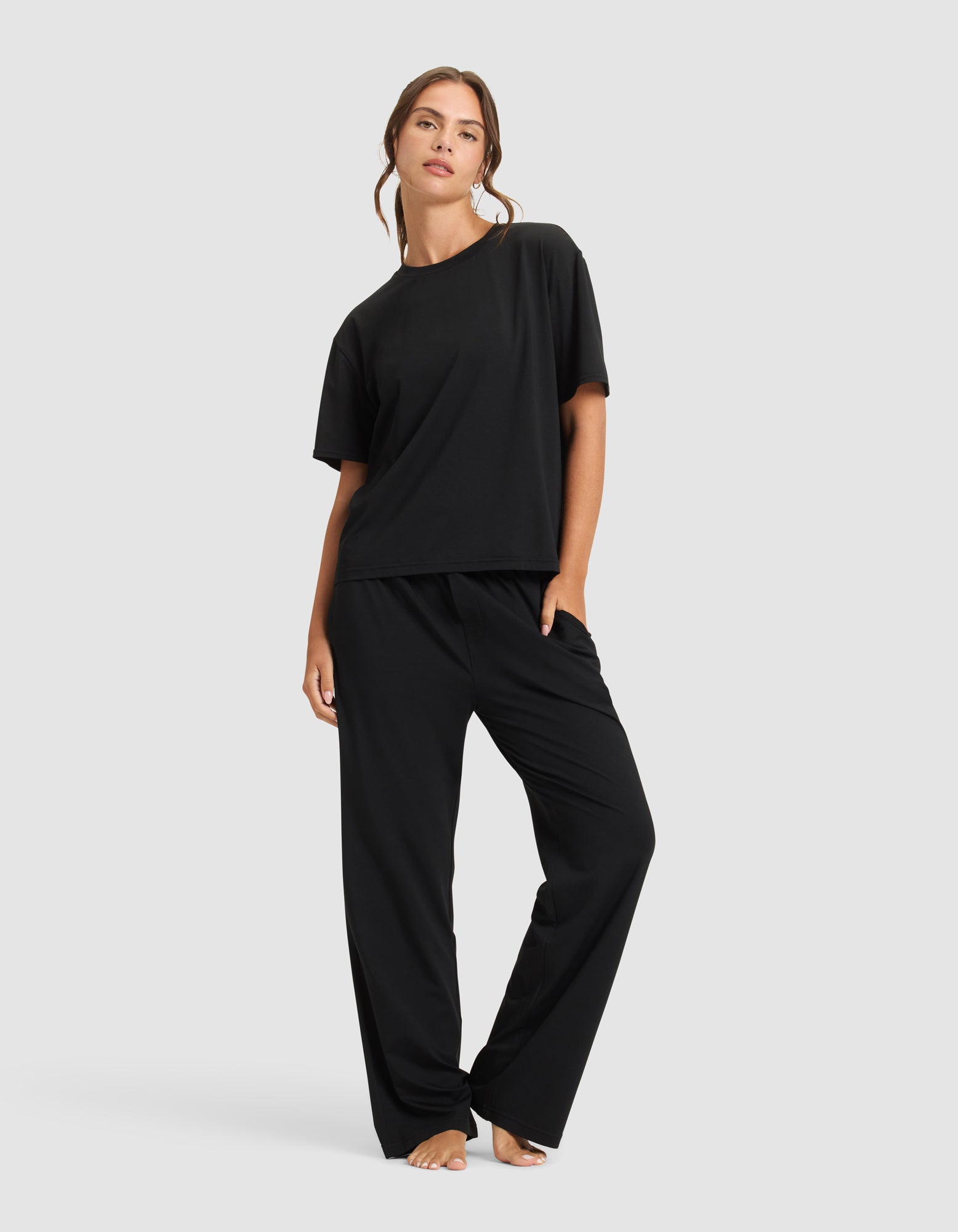 A woman poses barefoot against a plain background wearing the Cozy Earth Women's First Layer Pajama Short Sleeve Tee in black, paired with matching wide-leg pants, with one leg slightly bent and her head tilted.