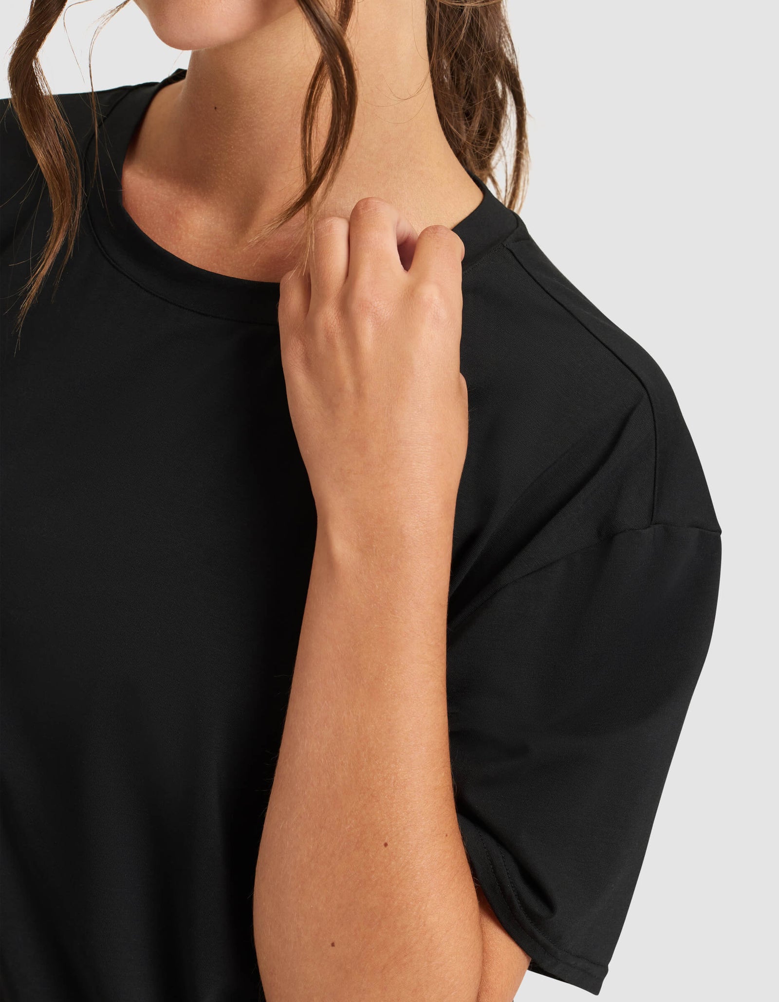 A woman wears the Cozy Earth Women's First Layer Pajama Short Sleeve Tee in black, with one hand raised near her shoulder. She is set against a light gray background, her face partially visible and brown hair loosely styled.