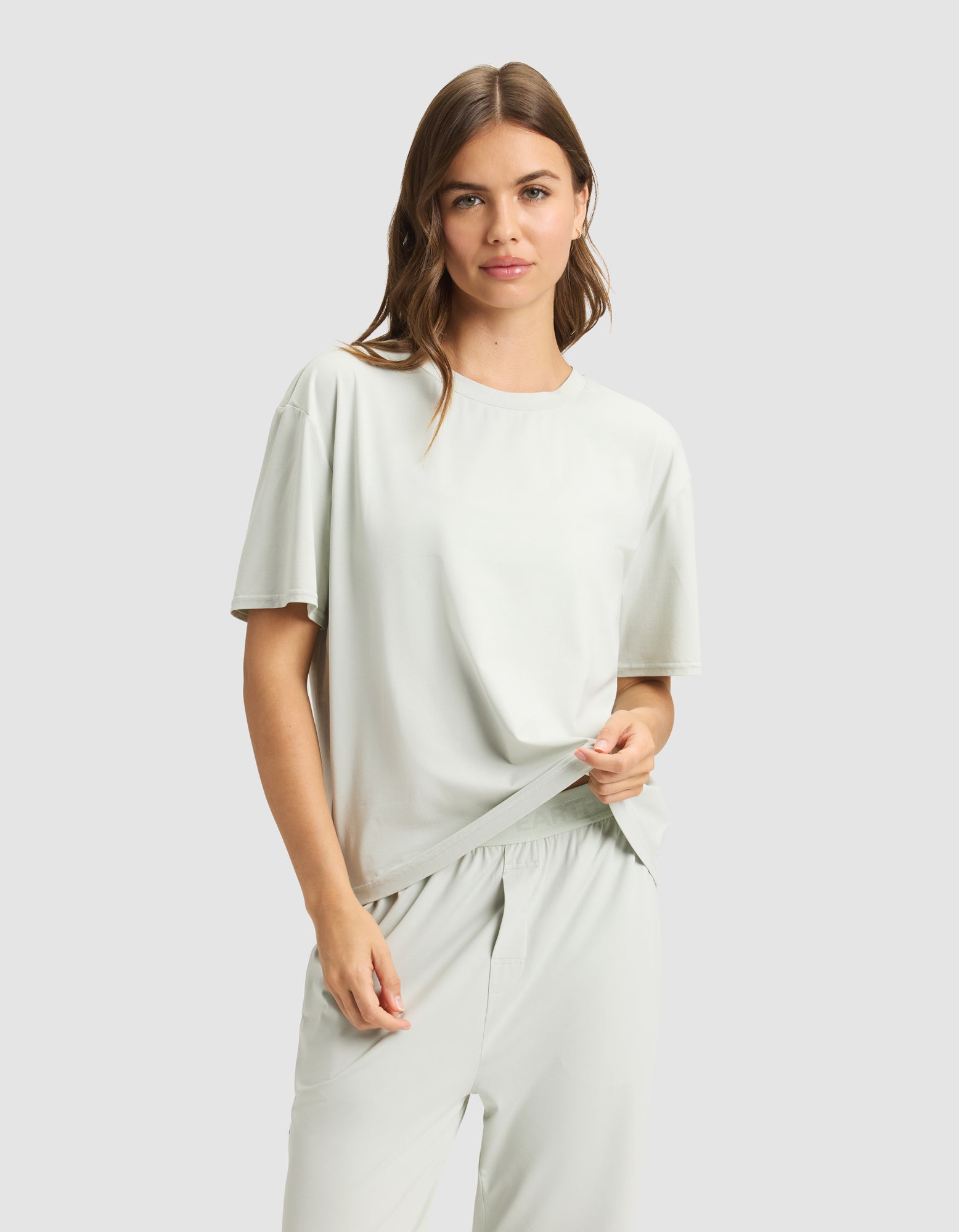 A woman with light brown hair wears the Cozy Earth Women's First Layer Pajama Short Sleeve Tee with matching pants, standing against a plain light gray background and smiling softly as she holds part of her shirt.