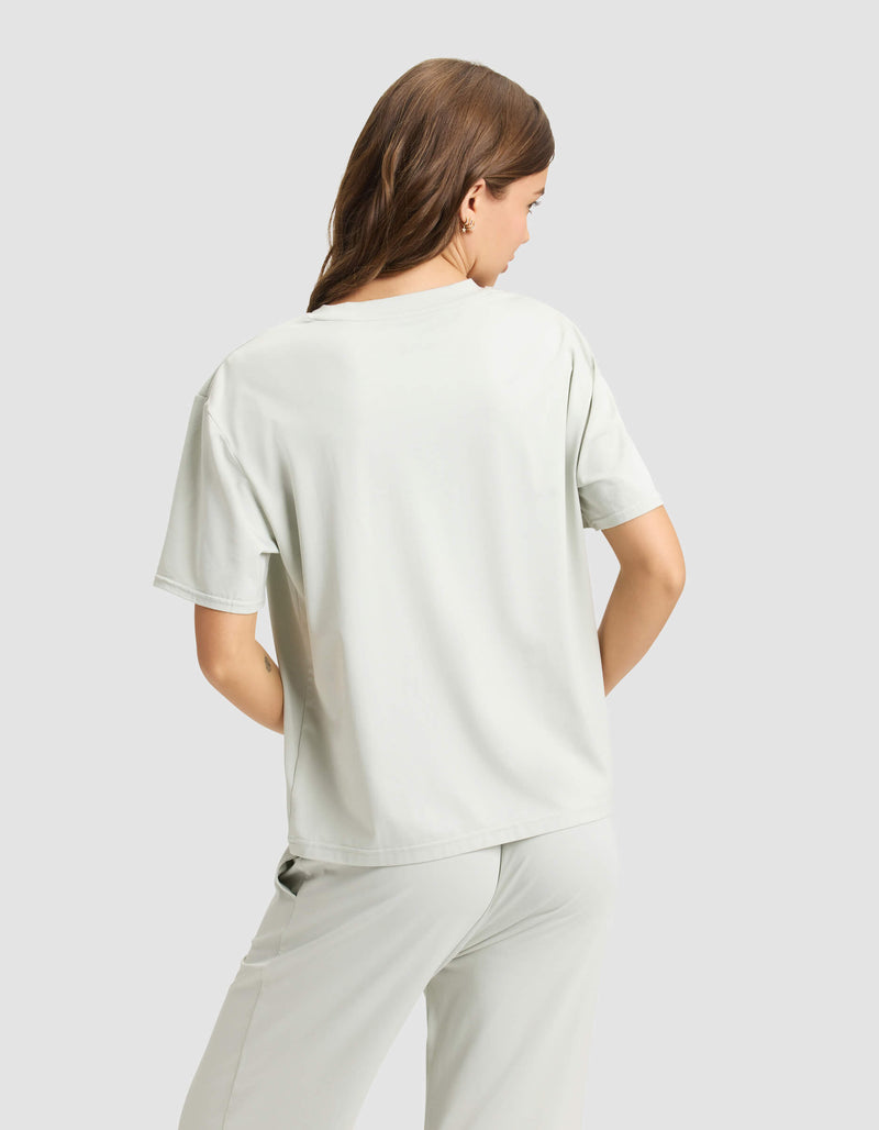 A woman with long brown hair stands with her back to the camera, wearing Cozy Earth's Women's First Layer Pajama Short Sleeve Tee in light gray, paired with matching pants against a plain light gray background.