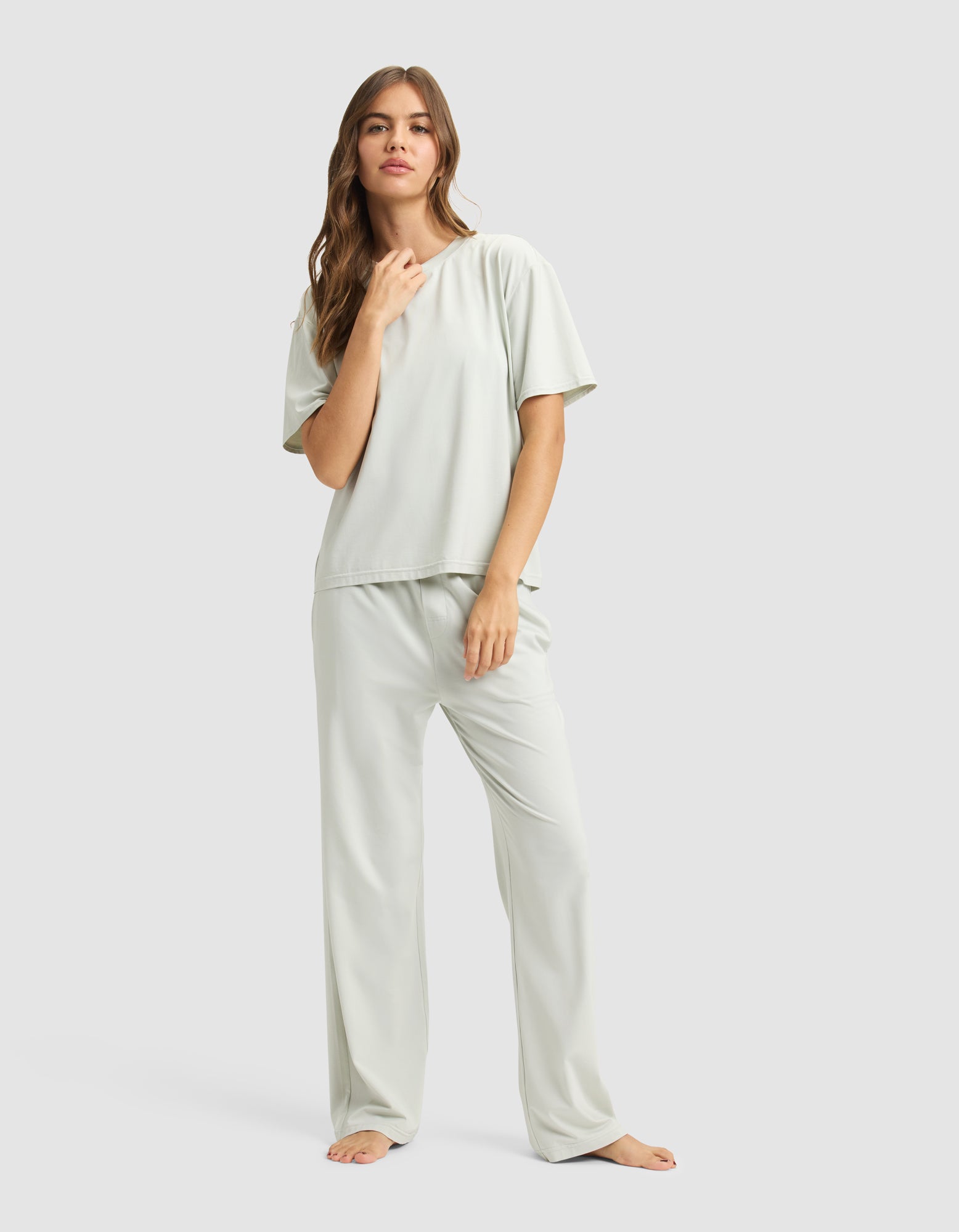 A woman with long, wavy hair stands barefoot against a plain background, confidently wearing Cozy Earth’s Women’s First Layer Pajama Short Sleeve Tee and matching pajama pants in a light color.