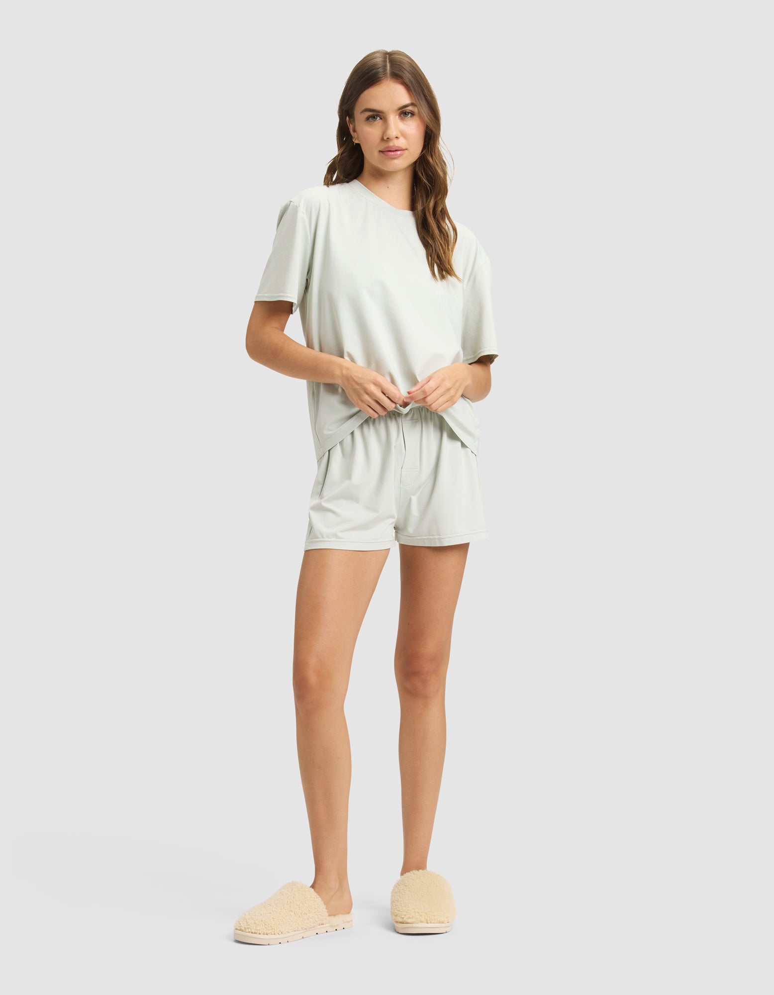 A woman with long wavy hair stands against a plain background wearing Cozy Earth's Women's First Layer Pajama Short Sleeve Tee in light sage green with matching shorts and beige fuzzy slippers, holding the waistband of her shorts.