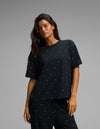 A woman with long dark hair stands against a plain gray background, wearing the Cozy Earth Women’s First Layer Pajama Short Sleeve Tee and matching black pants, both featuring small white polka dots.|Color:Mini Ghost