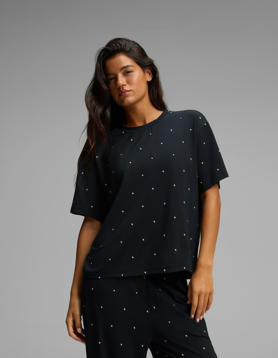A woman with long dark hair stands against a plain gray background, wearing the Cozy Earth Women’s First Layer Pajama Short Sleeve Tee and matching black pants, both featuring small white polka dots.
