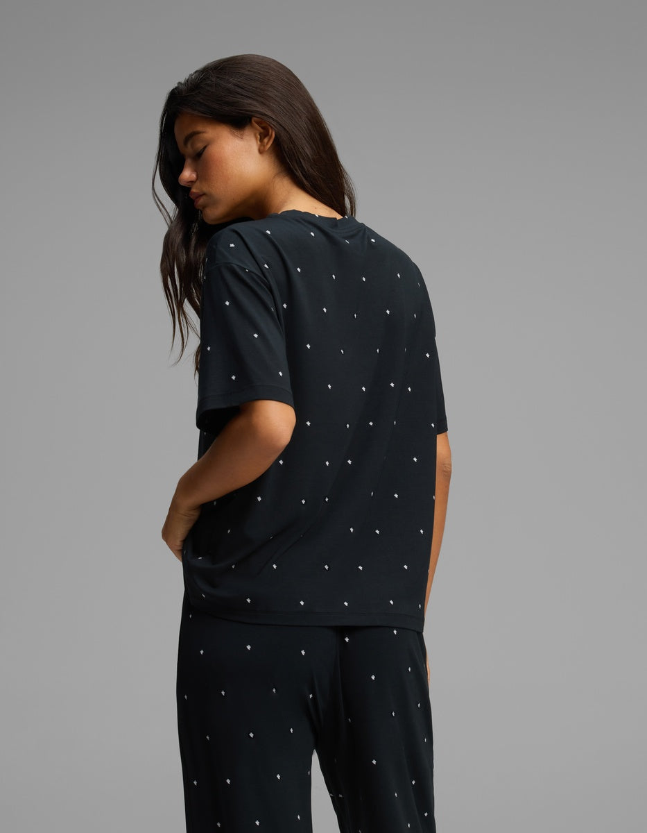 A woman with long brown hair stands sideways, looking down, wearing the Cozy Earth Women’s First Layer Pajama Short Sleeve Tee—a matching dark pajama set adorned with small white polka dots. Her right hand rests in her pocket against a plain light gray background.