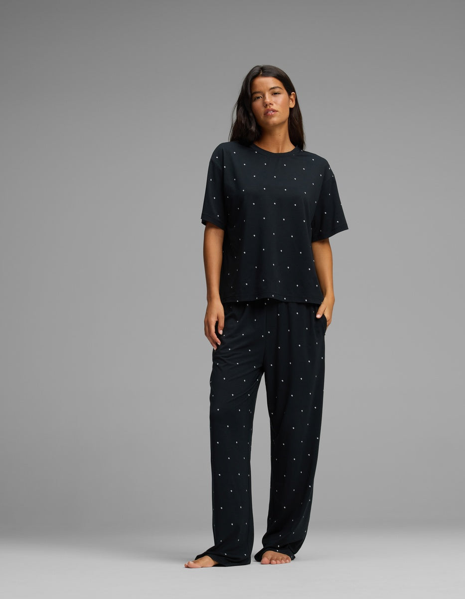 A woman stands barefoot against a plain gray background, wearing the Cozy Earth Women’s First Layer Pajama Short Sleeve Tee in a loose-fitting black set with small white polka dots.