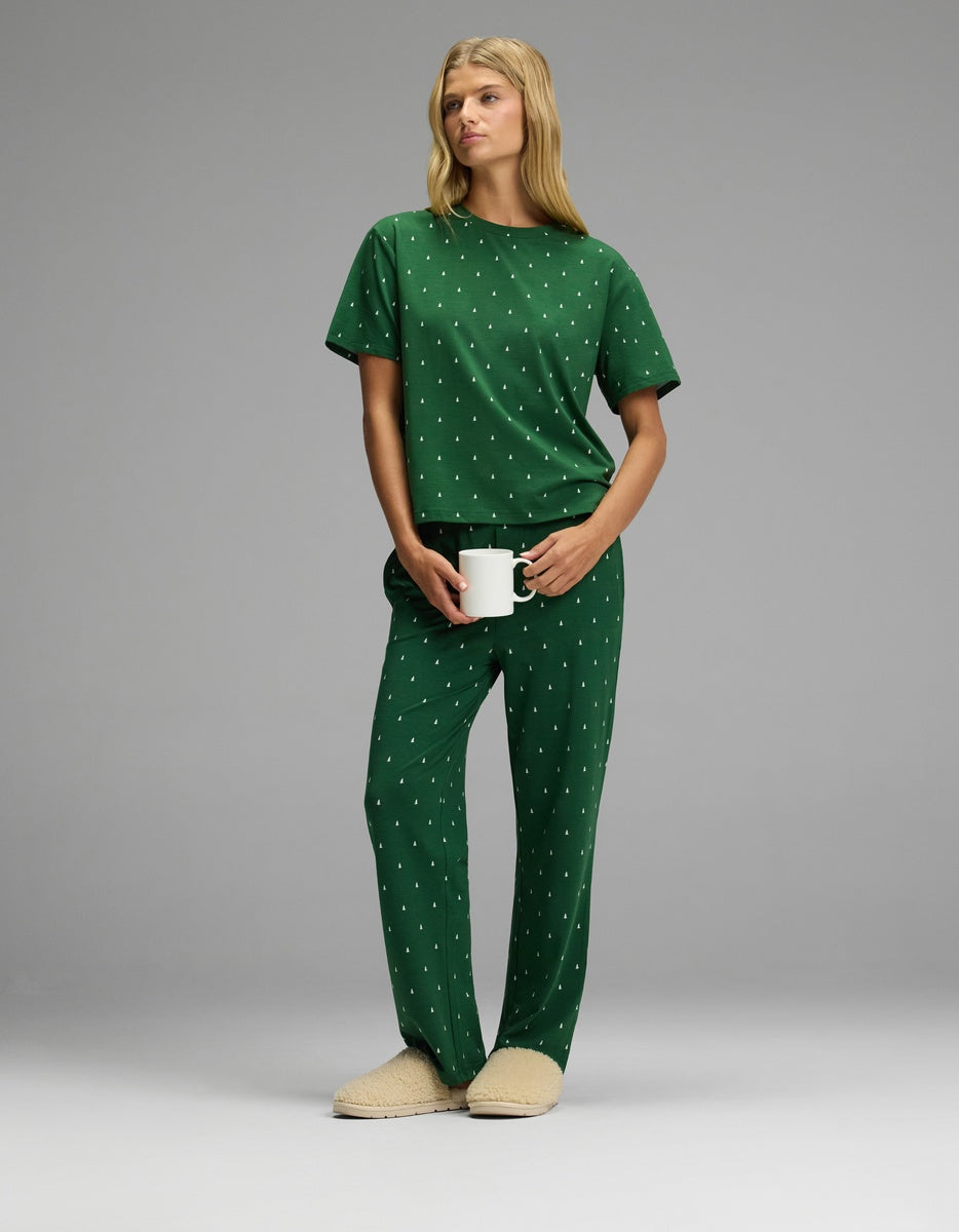 A woman wearing a green Women’s First Layer Pajama Short Sleeve Tee & Pajama Pant Set with a white pine tree print and beige slippers, holding a white mug.