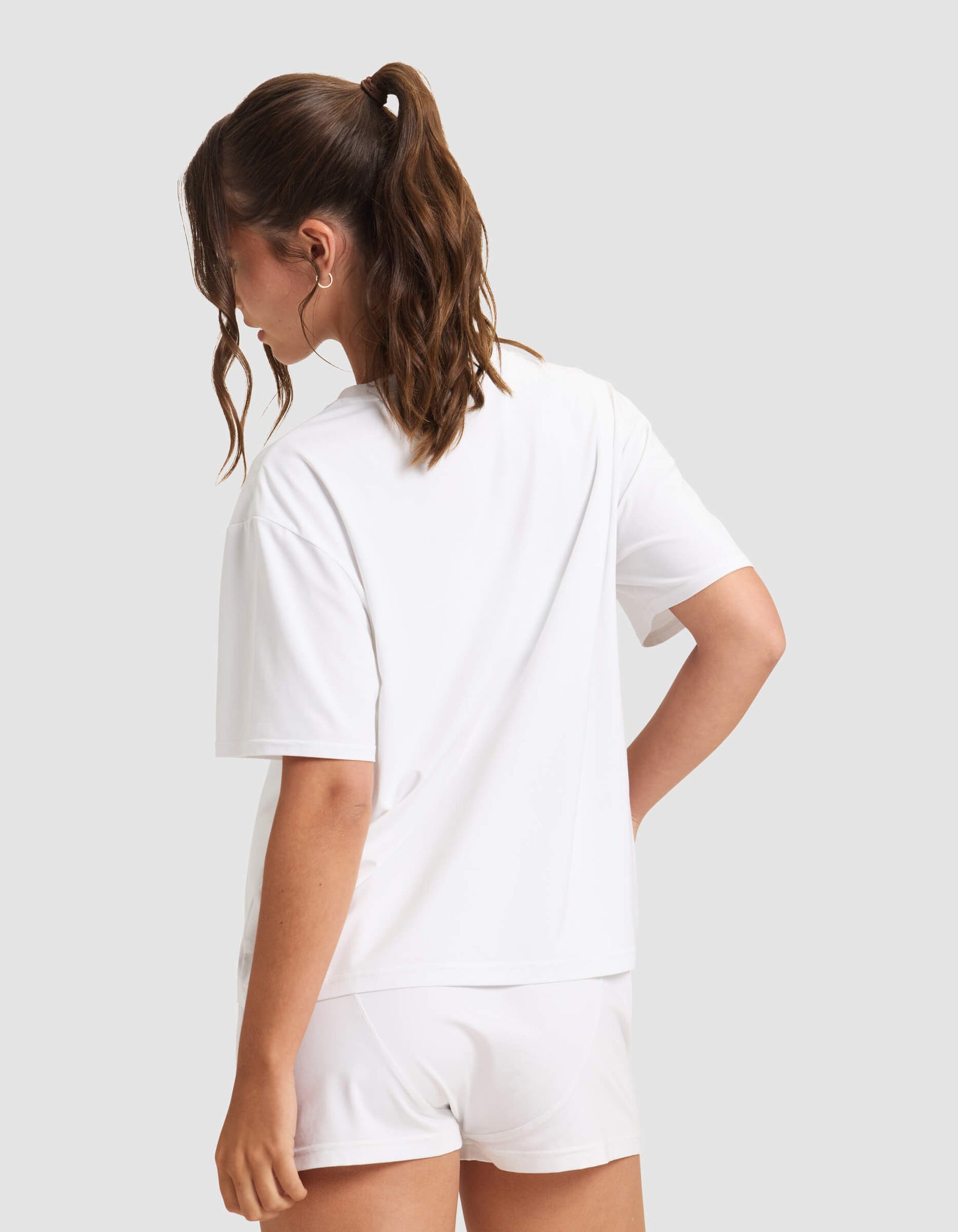 A woman with wavy brown hair in a ponytail stands with her back to the camera, wearing Cozy Earth's Women's First Layer Pajama Short Sleeve Tee and white shorts against a plain light gray background.