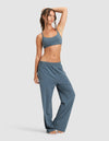 A woman poses barefoot against a plain background in the Cozy Earth Women's First Layer Pajama Soft Bra & Pant Set, featuring a matching blue sports bra and loose-fitting pajama pants, with one hand on her head and the other by her side.|Color:Dawn