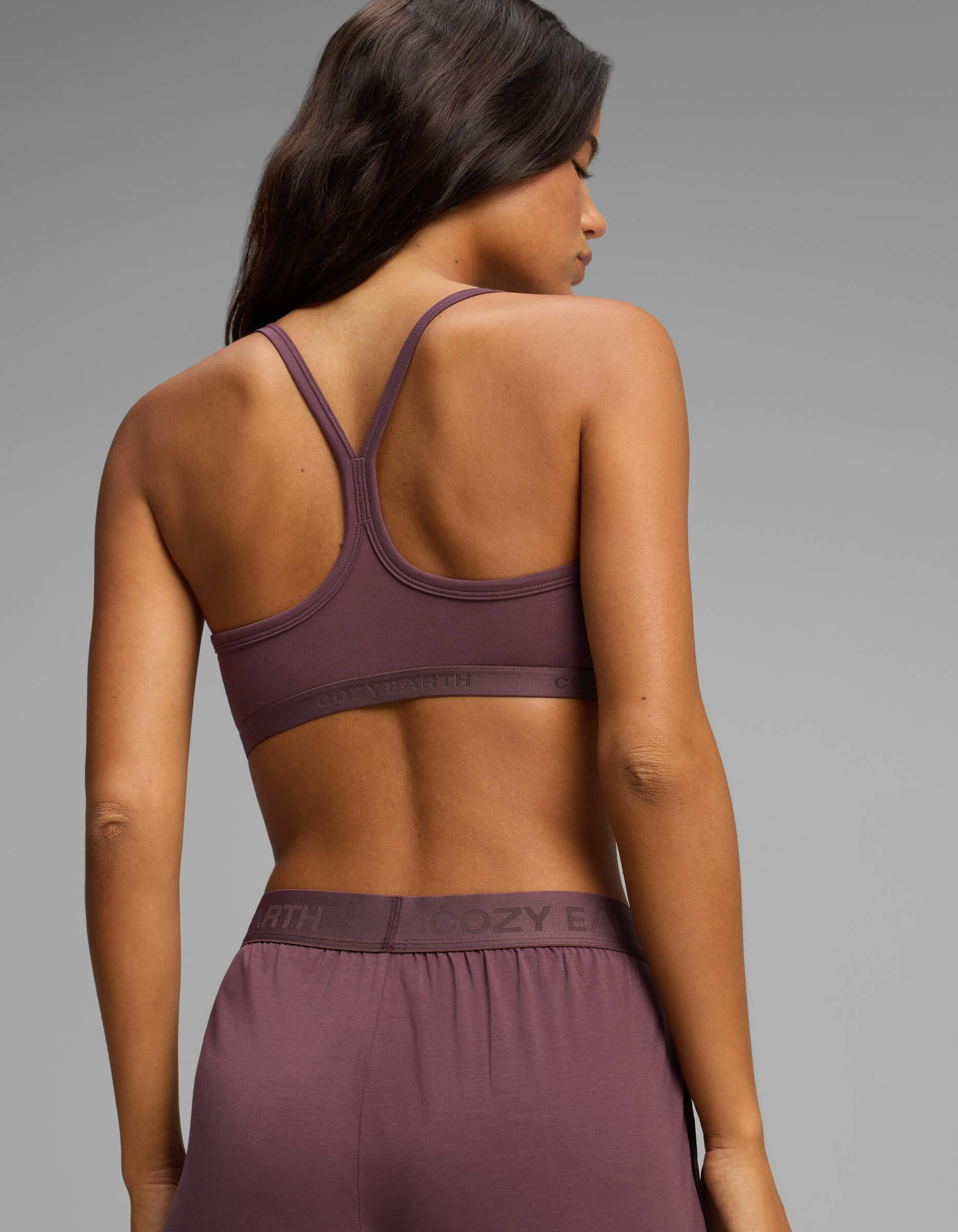A woman with long dark hair is shown from the back, wearing the Cozy Earth Women’s First Layer Soft Bra in mauve along with matching lounge pants, set against a plain gray background.