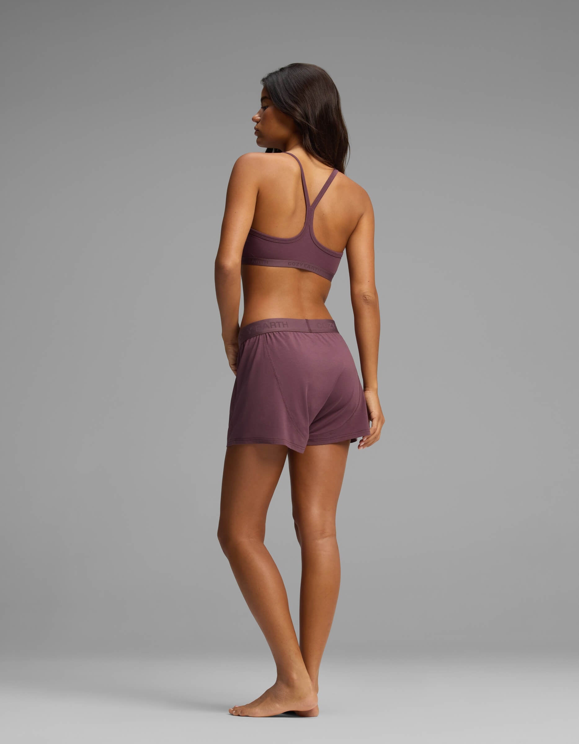 A woman stands barefoot against a plain gray background, facing away with her head turned to the right, wearing the Cozy Earth Women’s First Layer Pajama Soft Bra & Boxer Short Set.