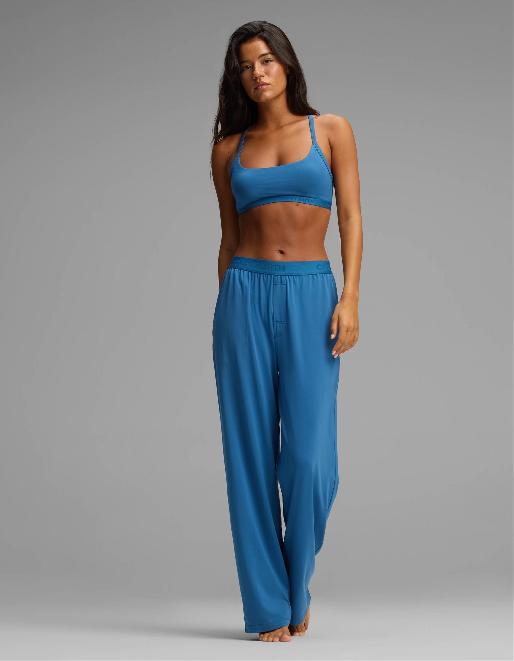 A woman stands barefoot in a studio, modeling the Cozy Earth Women’s First Layer Pajama Soft Bra & Pant Set in blue. She has long dark hair and a neutral expression, set against a plain gray background.