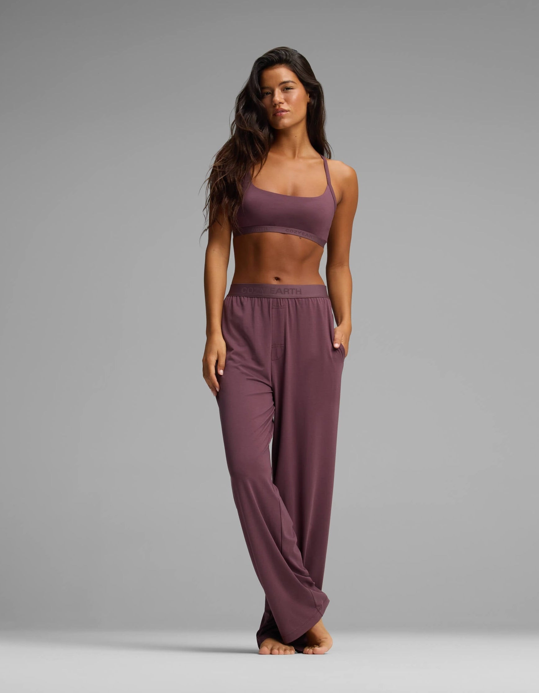 A woman stands barefoot on a gray background, wearing the Cozy Earth Women’s First Layer Pajama Soft Bra & Pant Set in mauve, with one hand in her pocket and the other by her side.