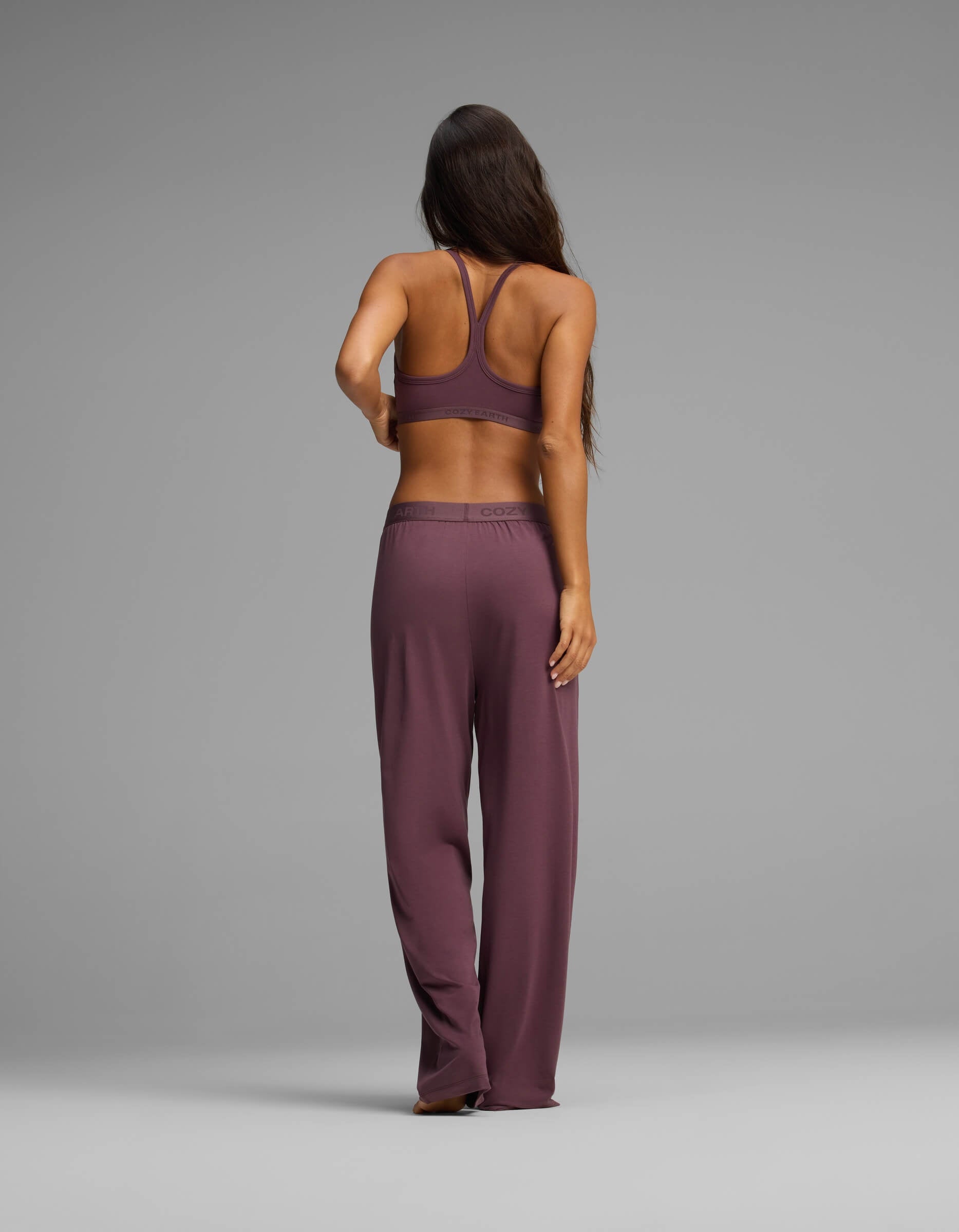 A woman with long brown hair, seen from behind, models the Cozy Earth Women’s First Layer Pajama Soft Bra & Pant Set in mauve, standing barefoot against a plain gray background.