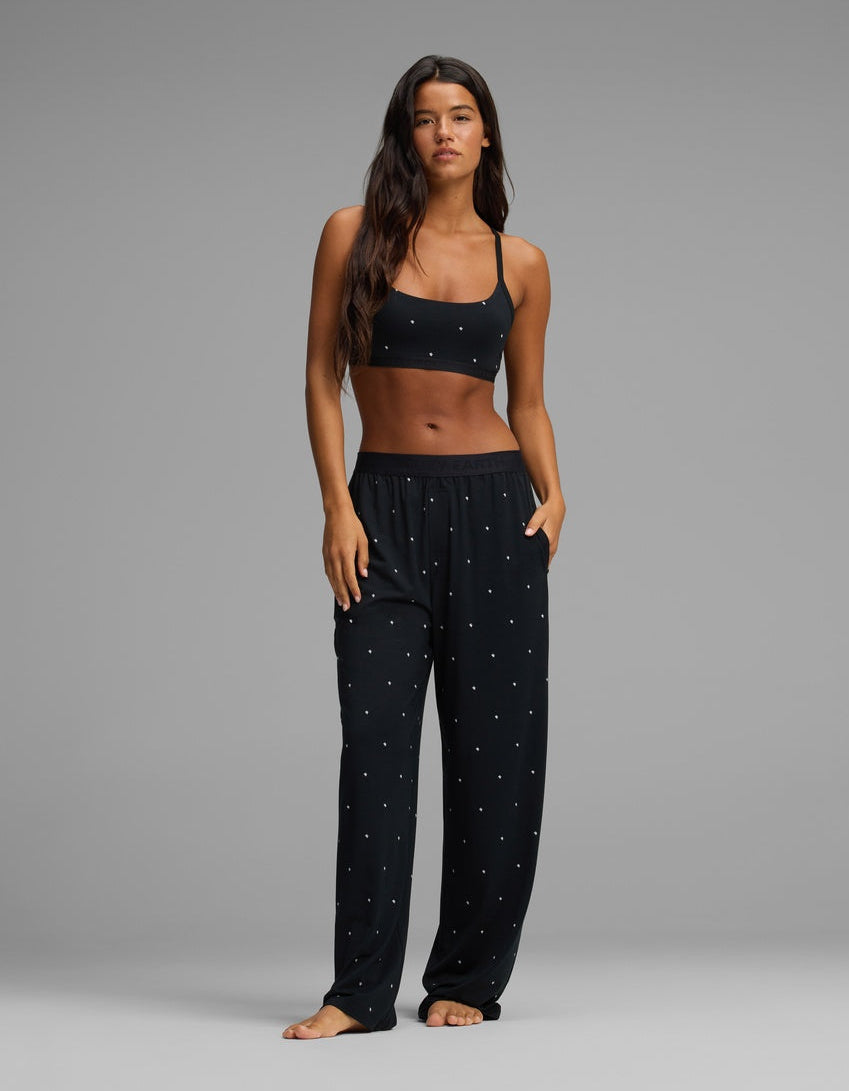 A woman stands barefoot against a plain gray background, wearing the Cozy Earth Women’s First Layer Pajama Soft Bra & Pajama Pant Set, featuring a black crop top and loose black pajama pants adorned with a small white dot pattern.|Color:Mini Ghost