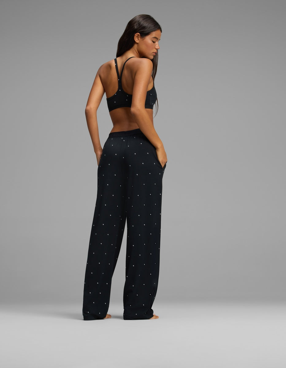 A woman stands facing away, wearing the Cozy Earth Women’s First Layer Pajama Soft Bra & Pajama Pant Set in black with small white dots. Her long hair is down, her hands rest in her pockets, and she is set against a plain gray background.|Color:Mini Ghost