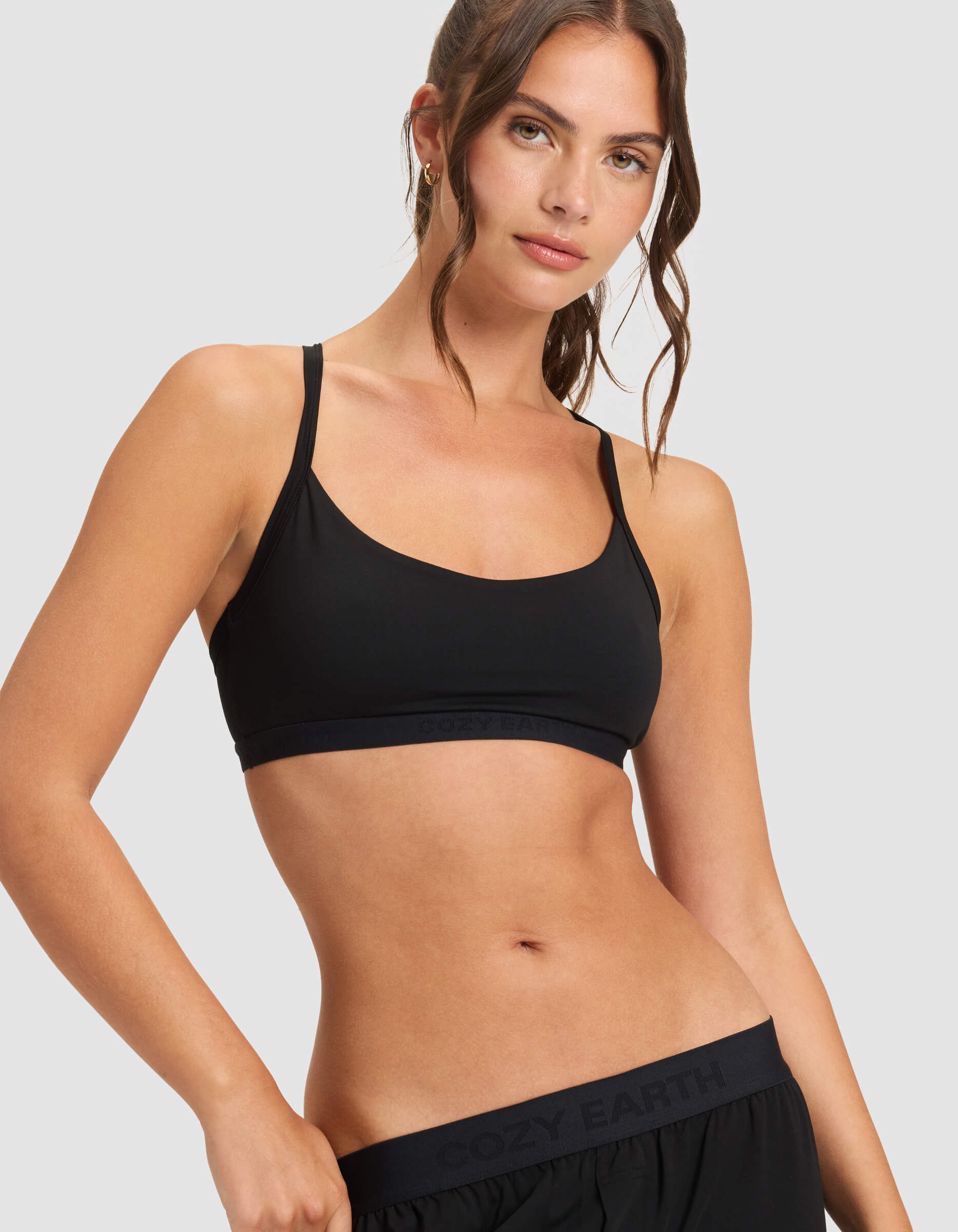 A woman with wavy brown hair wears the Cozy Earth Women's First Layer Pajama Soft Bra in black, paired with black shorts. She stands against a light gray background, looking slightly to the side with a relaxed, confident expression.|Color:Jet Black