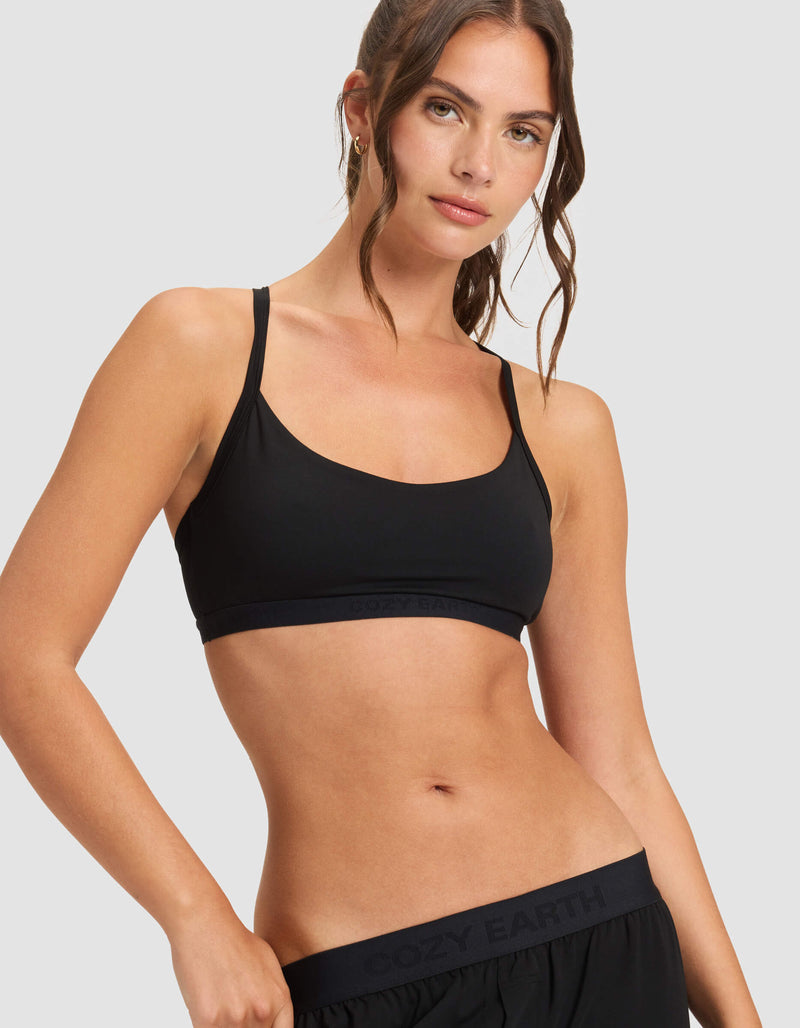 A woman with wavy brown hair wears the Cozy Earth Women's First Layer Pajama Soft Bra in black, paired with black shorts. She stands against a light gray background, looking slightly to the side with a relaxed, confident expression.