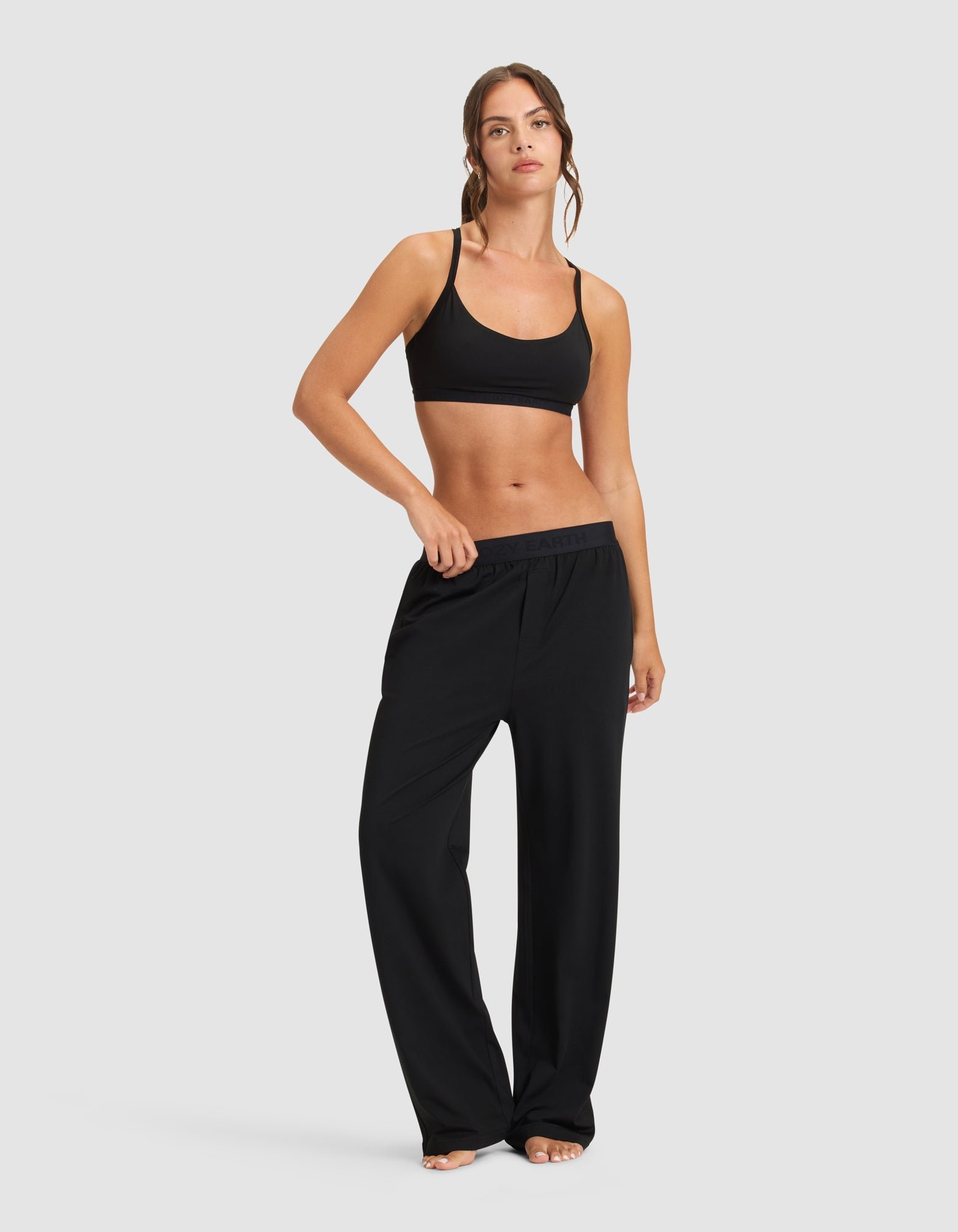 A woman stands barefoot against a plain backdrop, wearing Cozy Earth’s Women's First Layer PJ Soft Bra in black with loose black pants. She rests one hand on her waistband, looking relaxed.