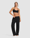 A woman stands barefoot against a plain backdrop, wearing Cozy Earth’s Women's First Layer PJ Soft Bra in black with loose black pants. She rests one hand on her waistband, looking relaxed.|Color:Jet Black