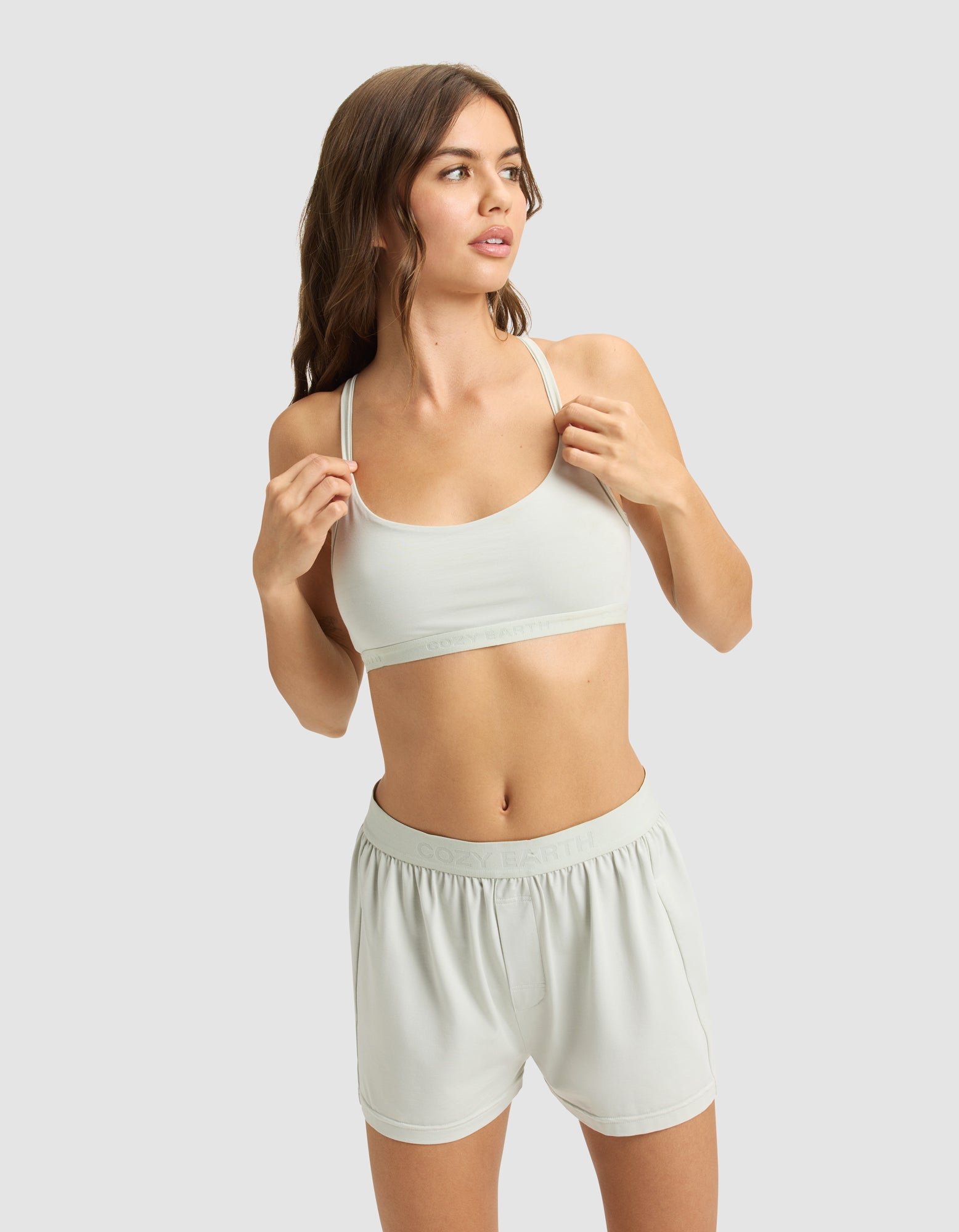 A woman with long brown hair models the Cozy Earth Women's First Layer PJ Soft Bra and matching shorts, standing against a plain light gray background and looking to the side.