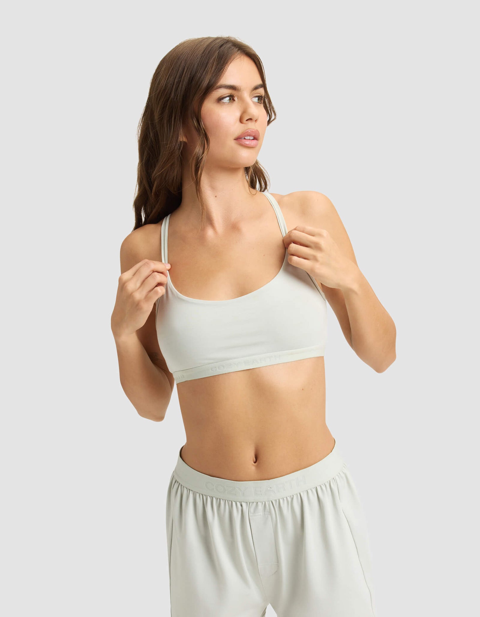 A woman with long brown hair models the Cozy Earth Women's First Layer PJ Soft Bra and matching shorts, standing against a plain light gray background and looking to the side.