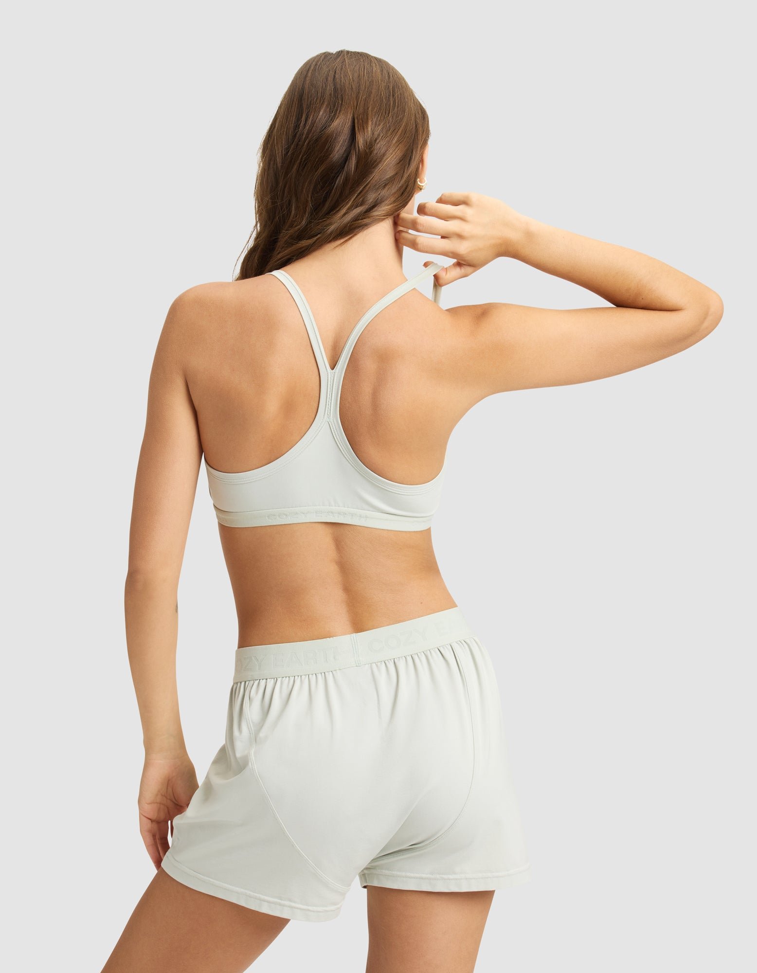 A woman with long brown hair faces away, wearing the Cozy Earth Women's First Layer PJ Soft Bra and matching shorts in light gray, standing against a plain light gray background with her right hand raised near her head.