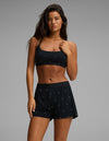 A woman wears the Cozy Earth Women’s First Layer Soft Bra in black, paired with matching black shorts featuring small white dots, as she stands against a plain gray background and looks directly at the camera.|Color:Mini Ghost