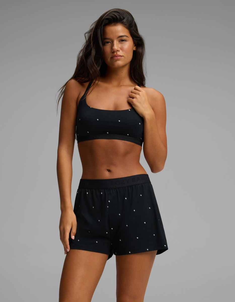 A woman wears the Cozy Earth Women’s First Layer Soft Bra in black, paired with matching black shorts featuring small white dots, as she stands against a plain gray background and looks directly at the camera.