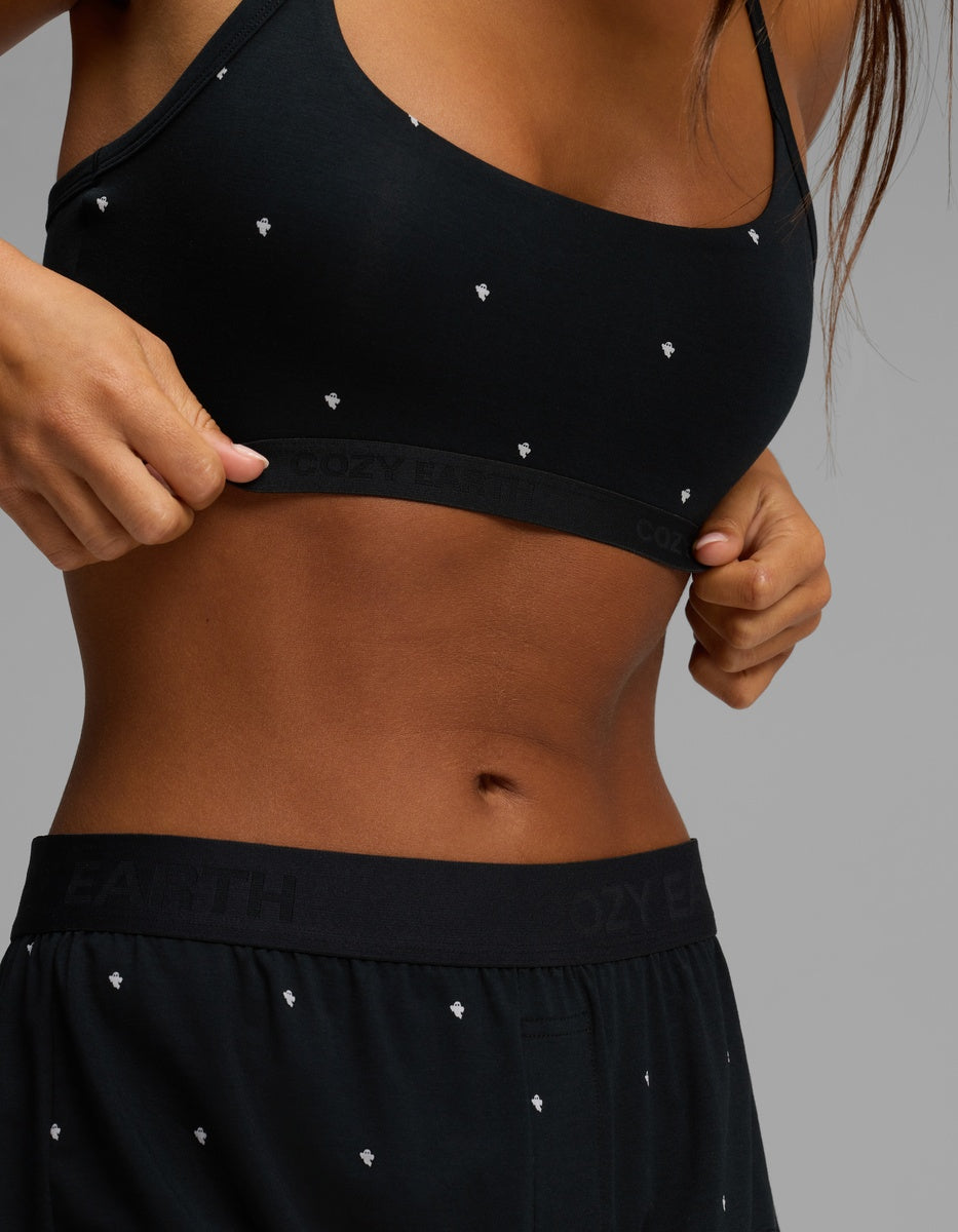 A woman wearing the Cozy Earth Women’s First Layer Soft Bra and matching black shorts with small white patterns adjusts her top, revealing part of her midriff against a neutral background.