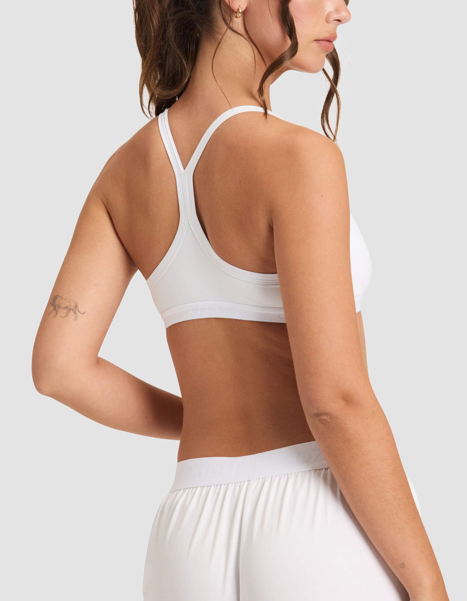 A woman is shown from the back wearing the Cozy Earth Women's First Layer Pajama Soft Bra in white with matching shorts, her hand on her hip and a small tattoo visible on her upper left arm.