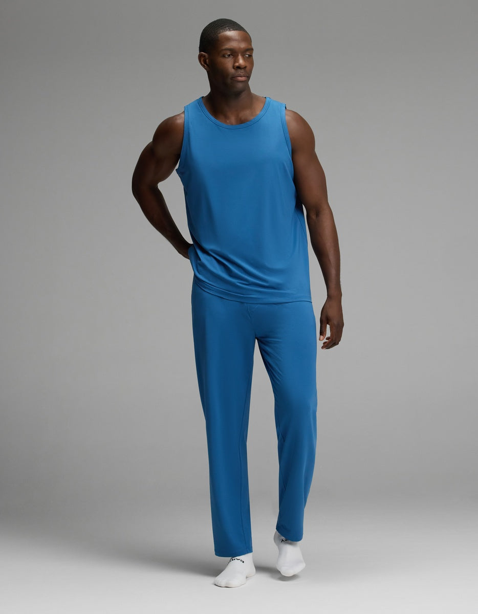 A man models the Cozy Earth Men’s First Layer Pajama Tank & Pant Set in blue, standing on a gray background with one hand resting on his hip and looking to the side. He completes the look with white socks.